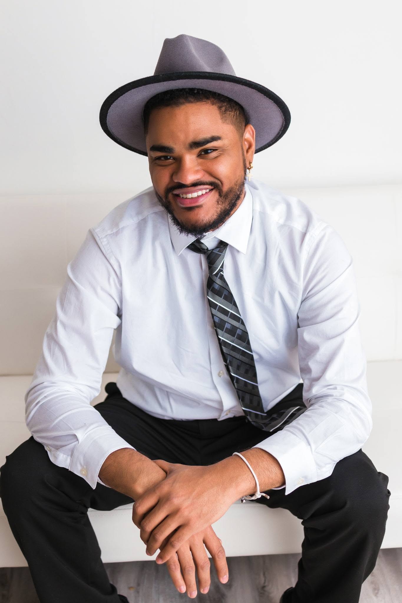Man with a beard wearing a hat, white shirt, and tie, sitting and smiling.