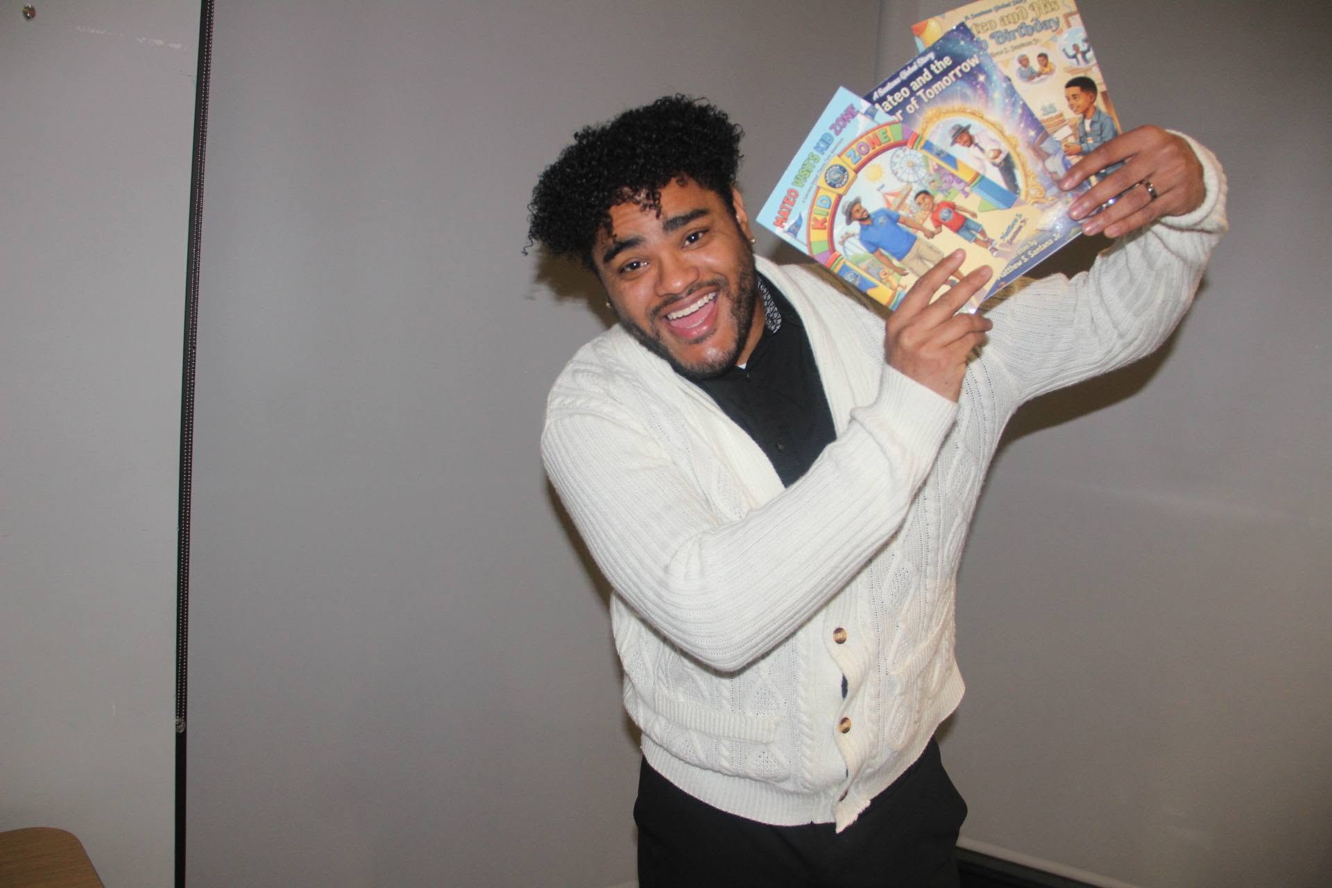 Person with curly hair smiling and holding colorful books or magazines, wearing a white cardigan, standing indoors.
