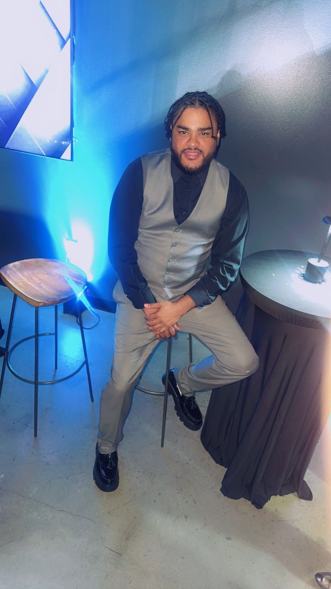 Man with glasses and beard sitting on a stool, smiling, in a room with blue lighting and a round table.