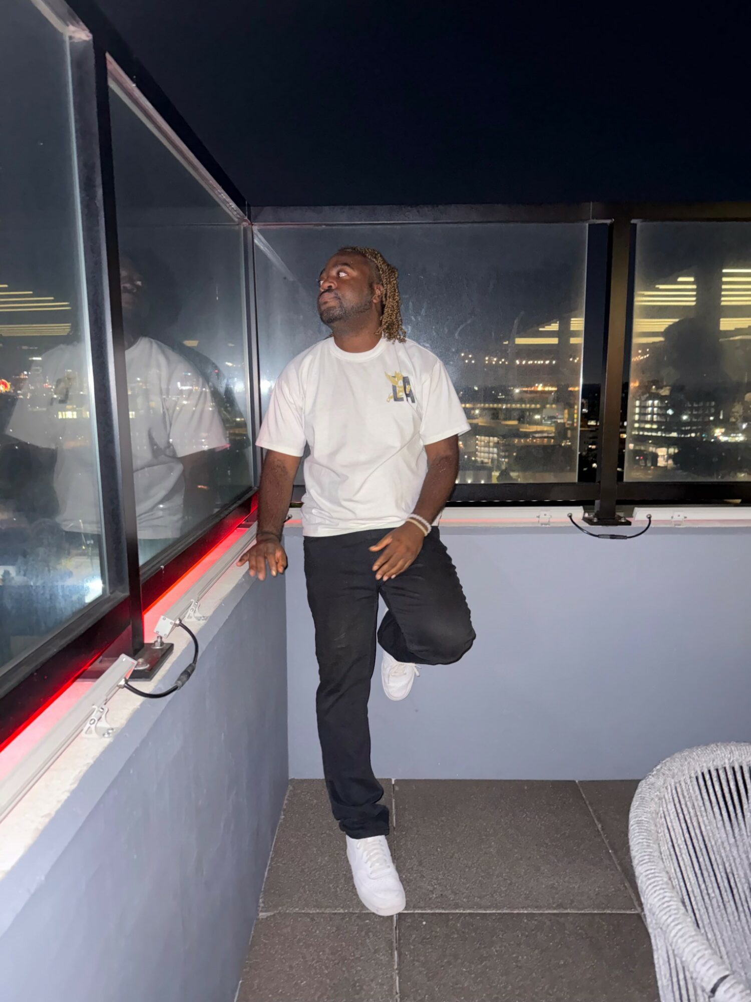 Person with blonde hair wearing a white t-shirt, black pants, and white shoes standing on a balcony at night, looking up.