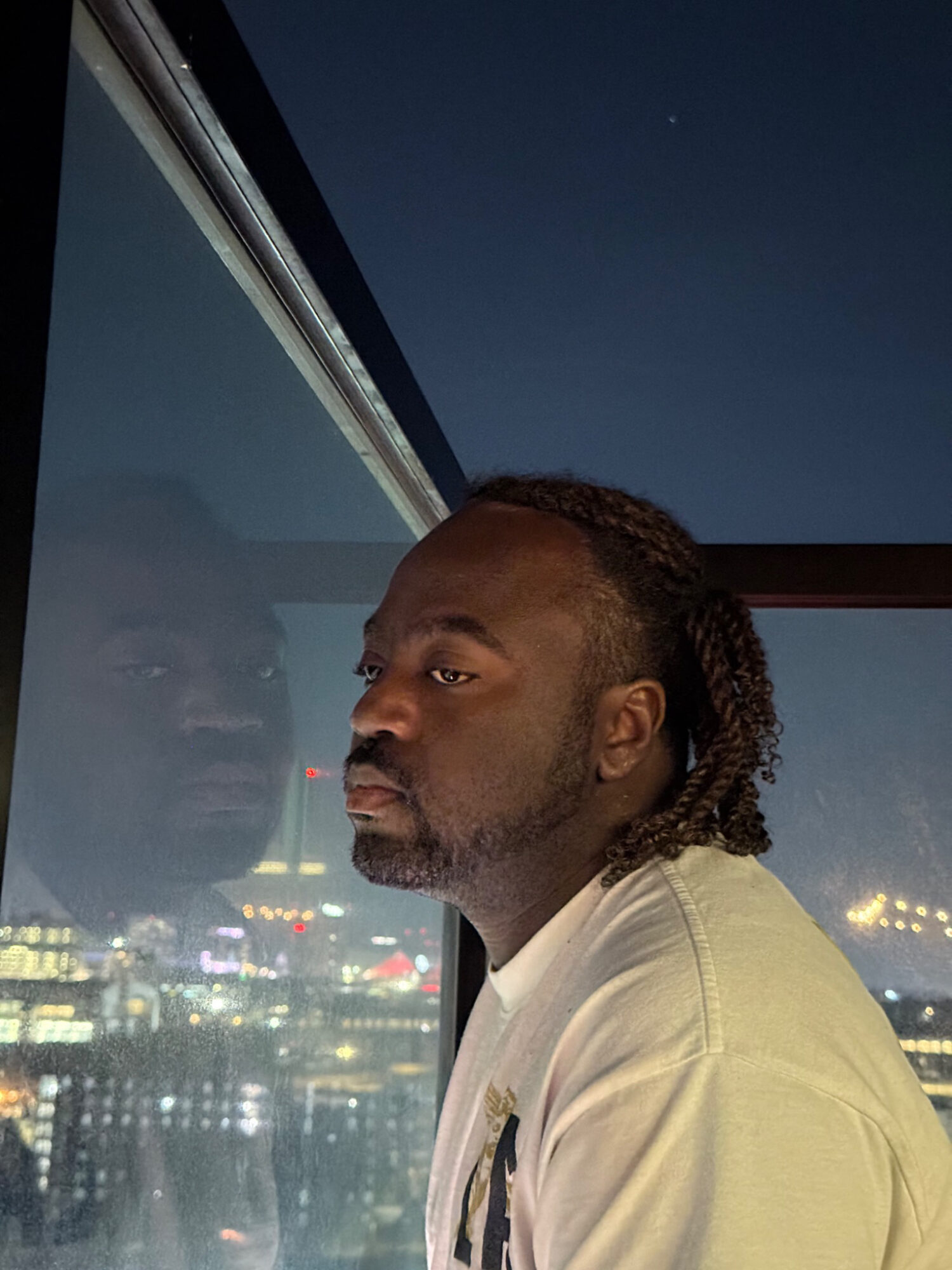 Man with dreadlocks looking out a window at night city lights, reflection visible in glass.