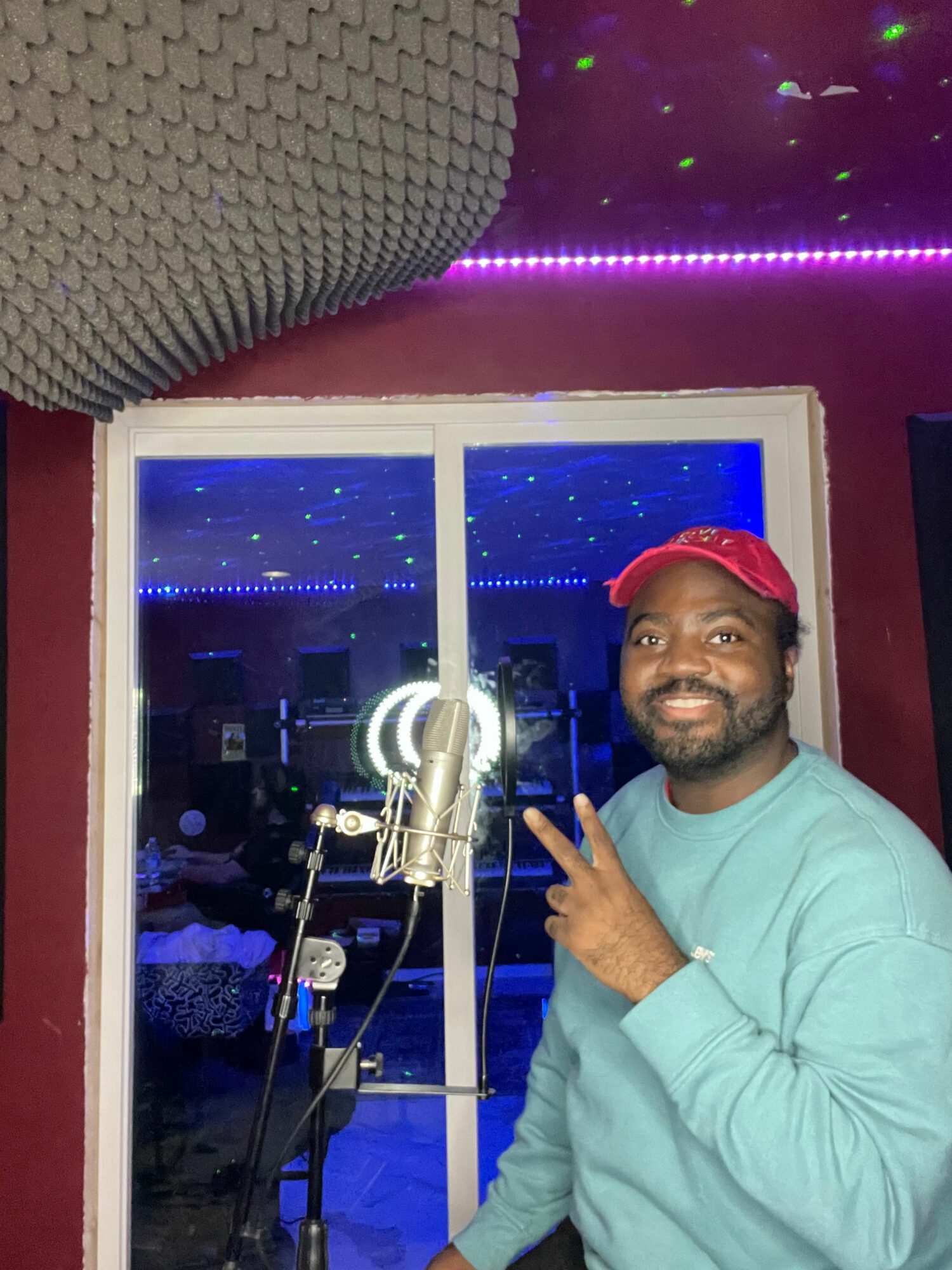 Man with a red cap smiling, pointing at a microphone in a recording studio with purple and blue lighting