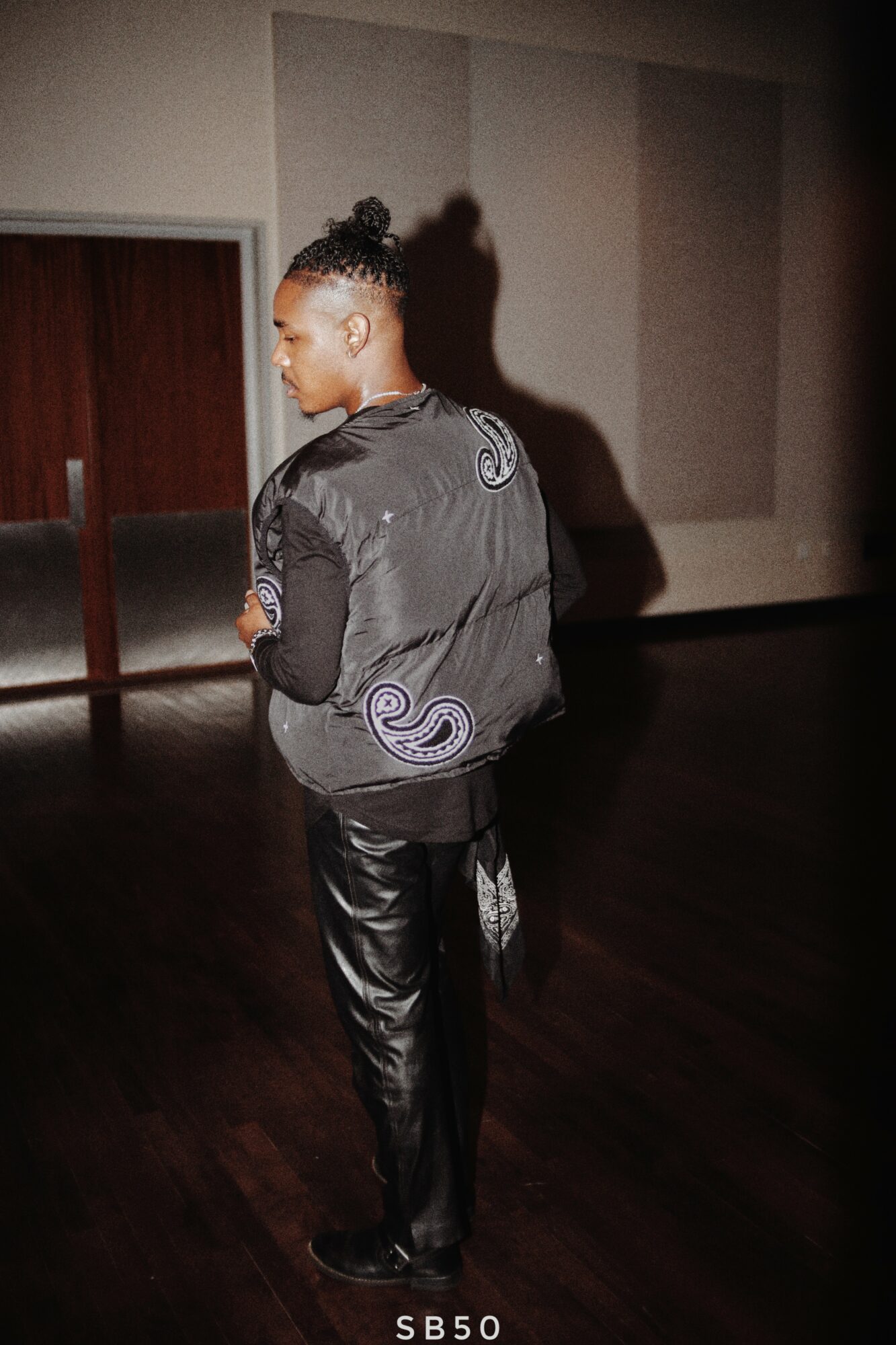 Person with braided hair wearing a black jacket and shiny pants, standing indoors with a shadow on the wall.