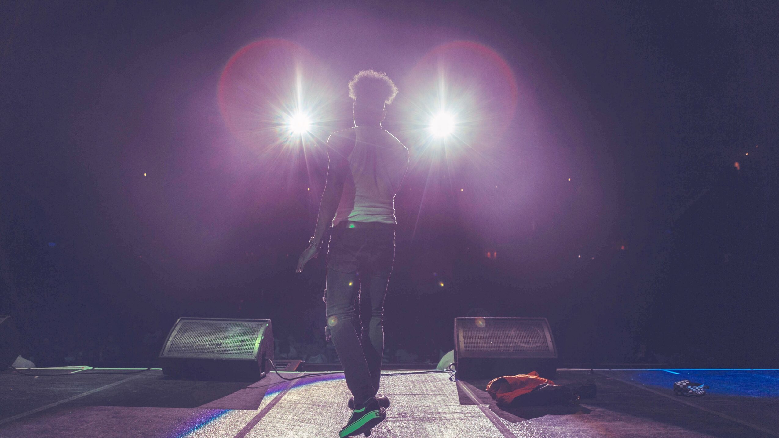 Person walking on stage with bright lights behind, dark background, and equipment on stage floor.