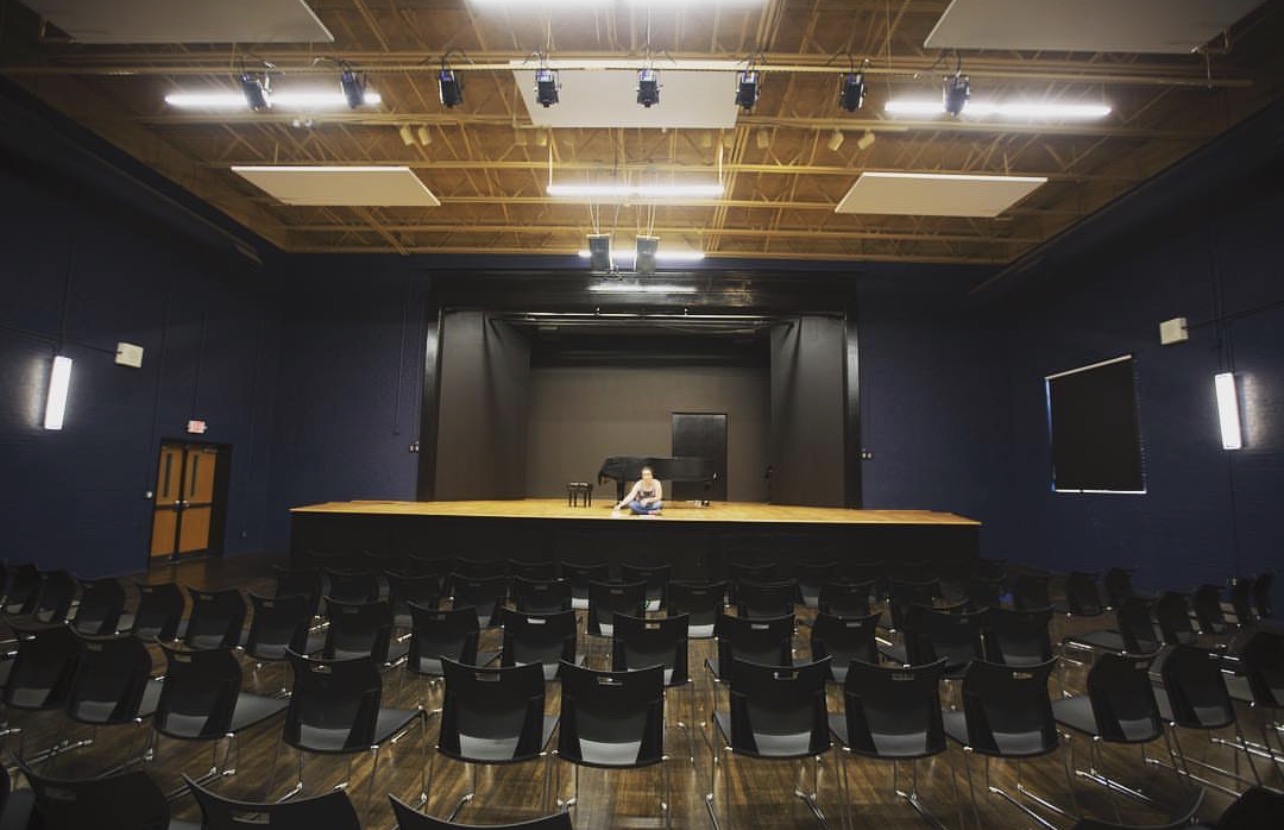 Empty auditorium with a stage, rows of chairs, and a person sitting at a desk on stage, ceiling lights overhead.