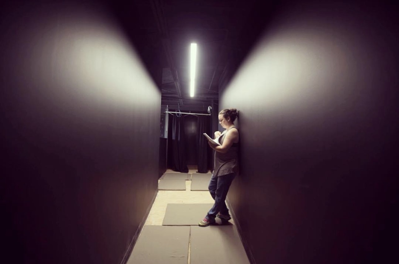 Person standing in a narrow corridor, looking at a phone, with dark walls and a bright ceiling light.