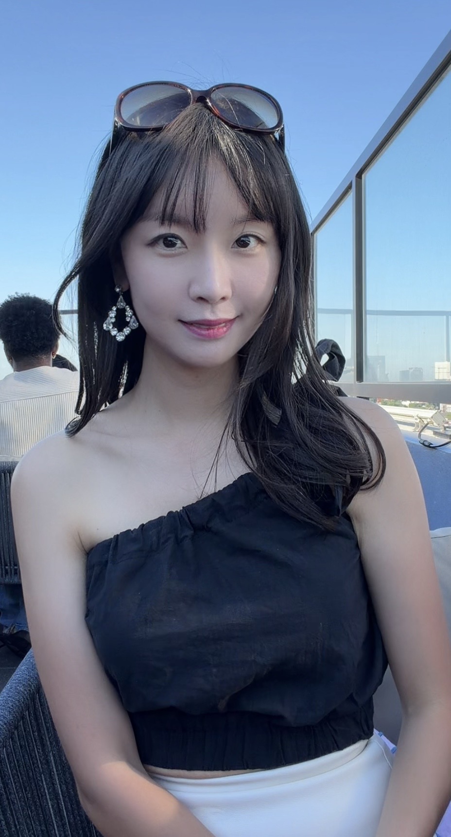 Young woman with sunglasses on her head, wearing a black off-shoulder top, smiling outdoors on a sunny day.