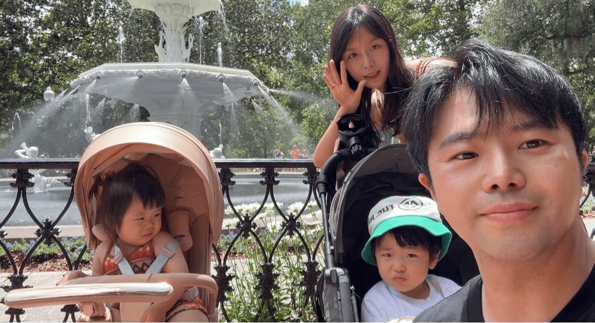 Family with two children in strollers outdoors near a fountain, smiling and posing for a photo.