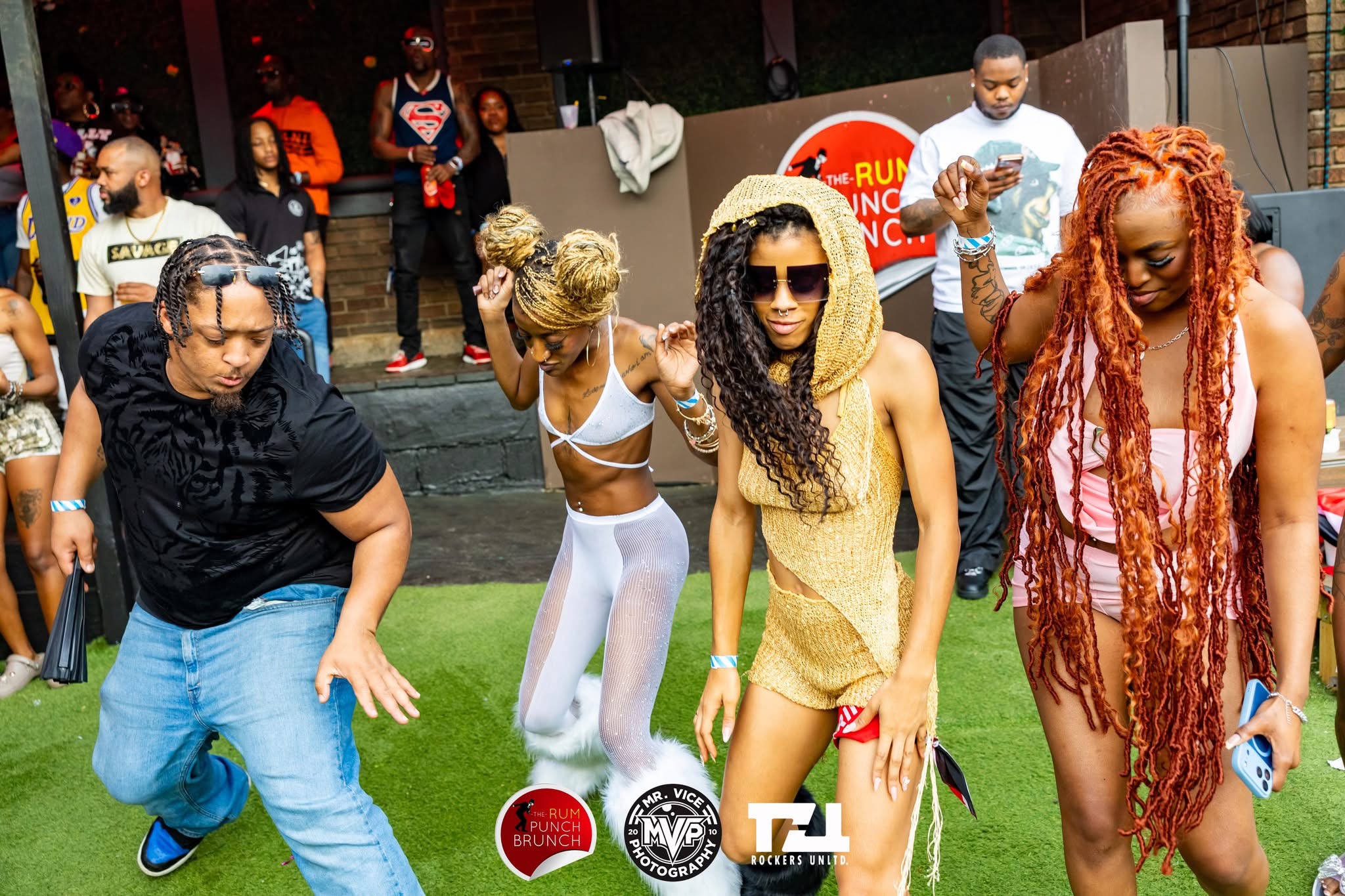 Group of four women dancing outdoors, some wearing sunglasses, with people in the background. Bright, lively scene.