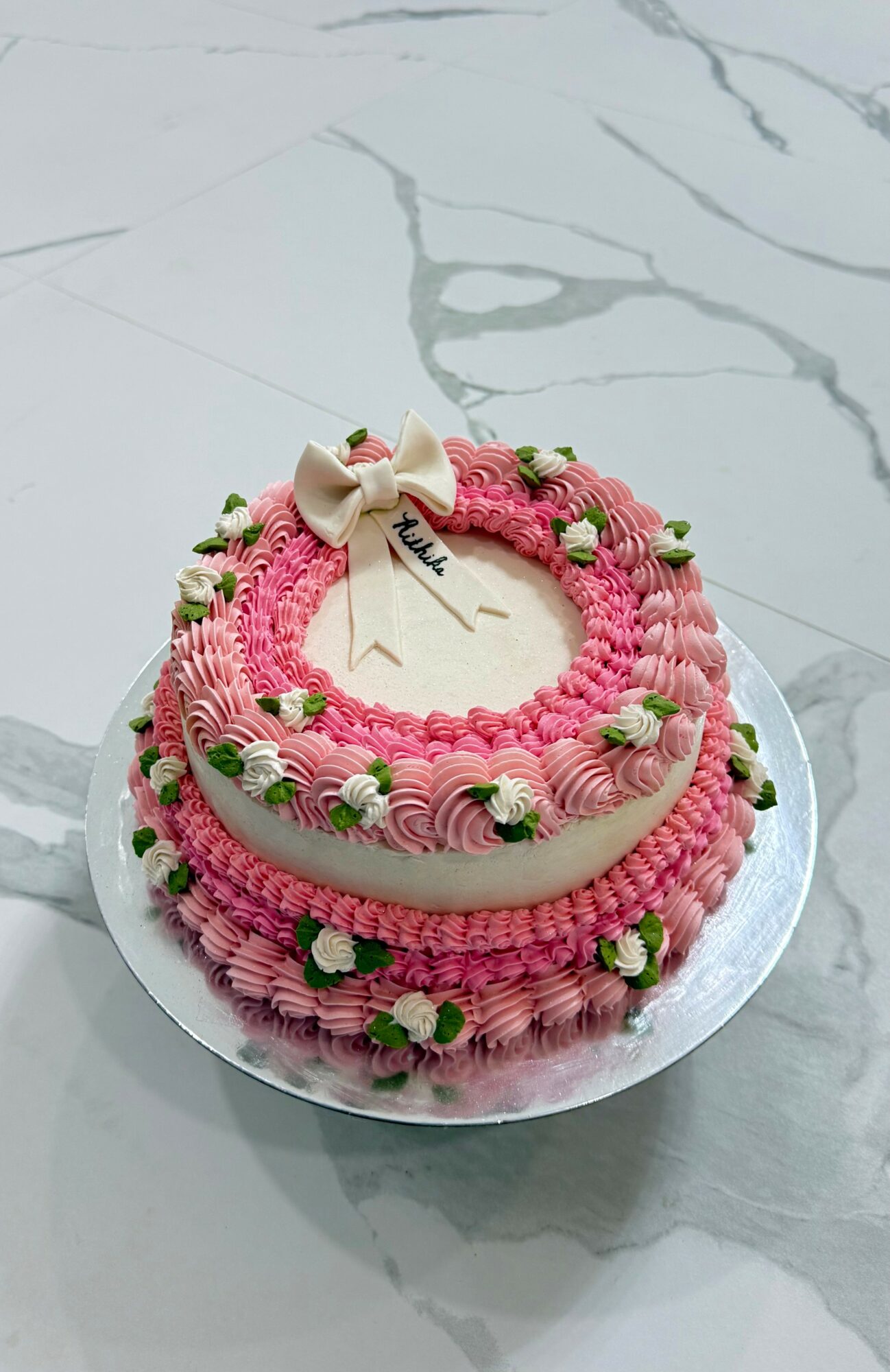 Heart-shaped cake decorated with pink and white frosting, green accents, and a bow on top, placed on a white marble surface.