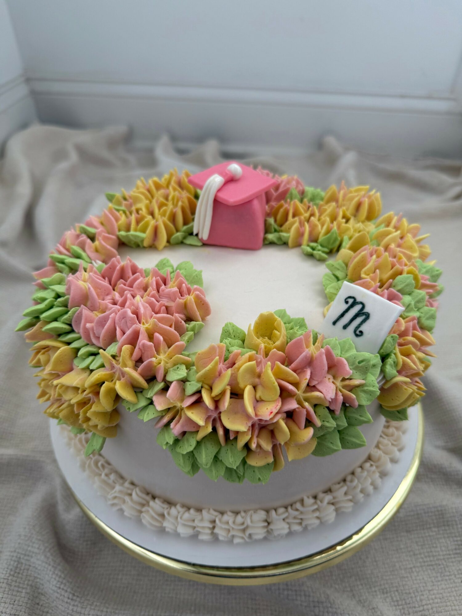Round cake decorated with colorful frosting flowers, a pink purse, and a white card with a black symbol, on a gold stand.