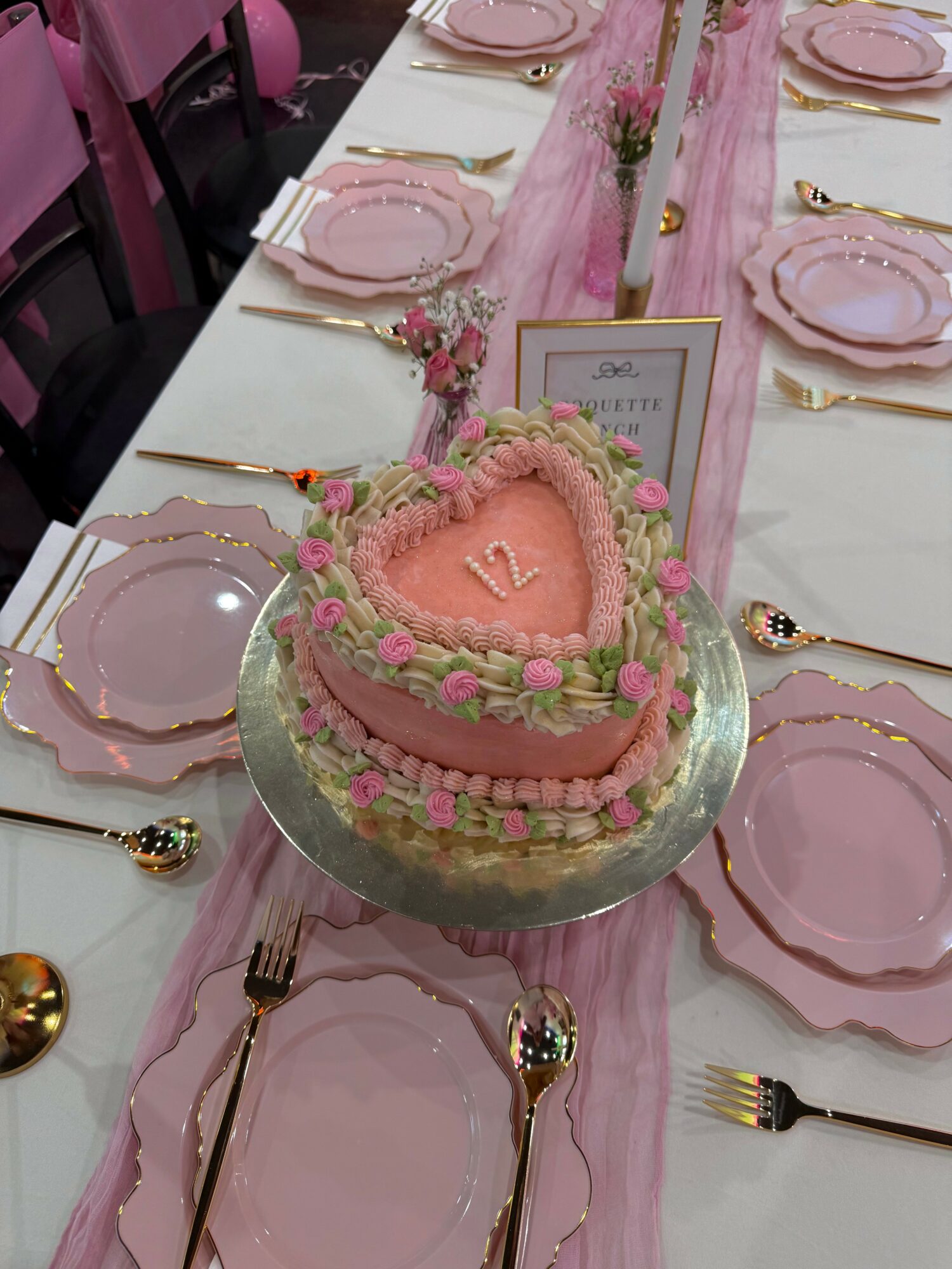 Table setting with pink plates, gold utensils, and a heart-shaped pink cake with green decorations, on a pink table runner.