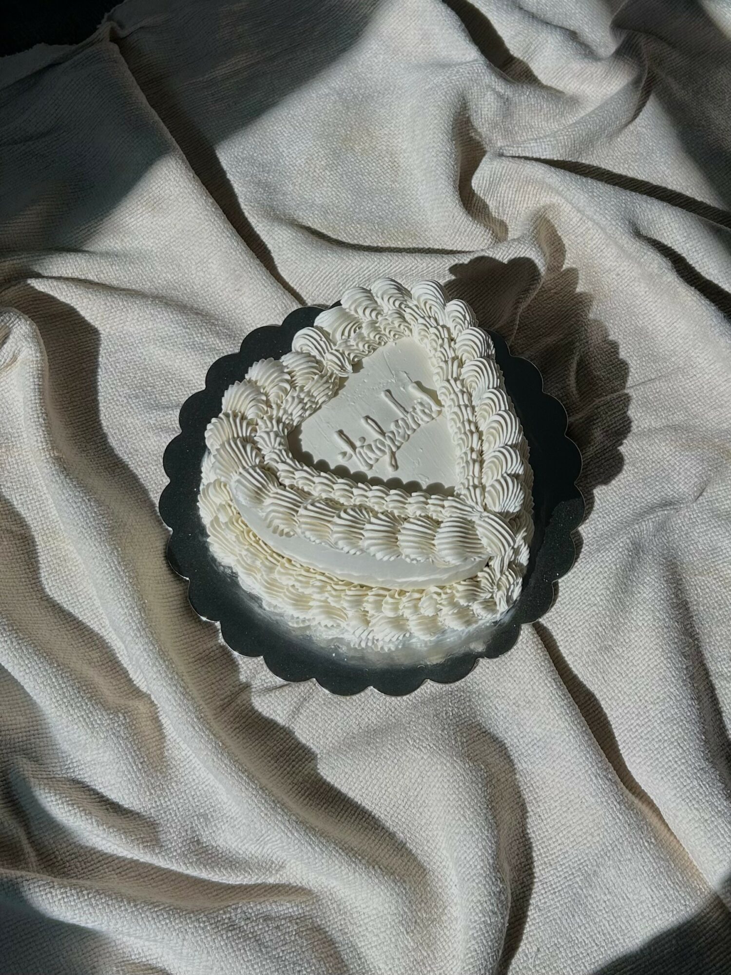 Heart-shaped cake with white frosting and decorative piping, placed on a black scalloped tray on a light fabric surface.