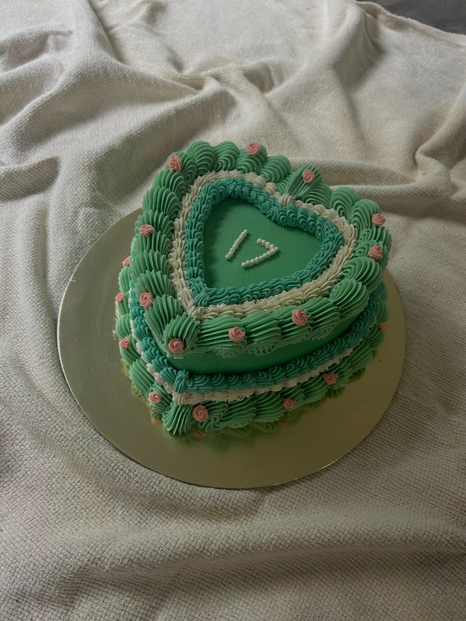 Heart-shaped cake decorated with green and pink icing, placed on a round silver board on fabric.