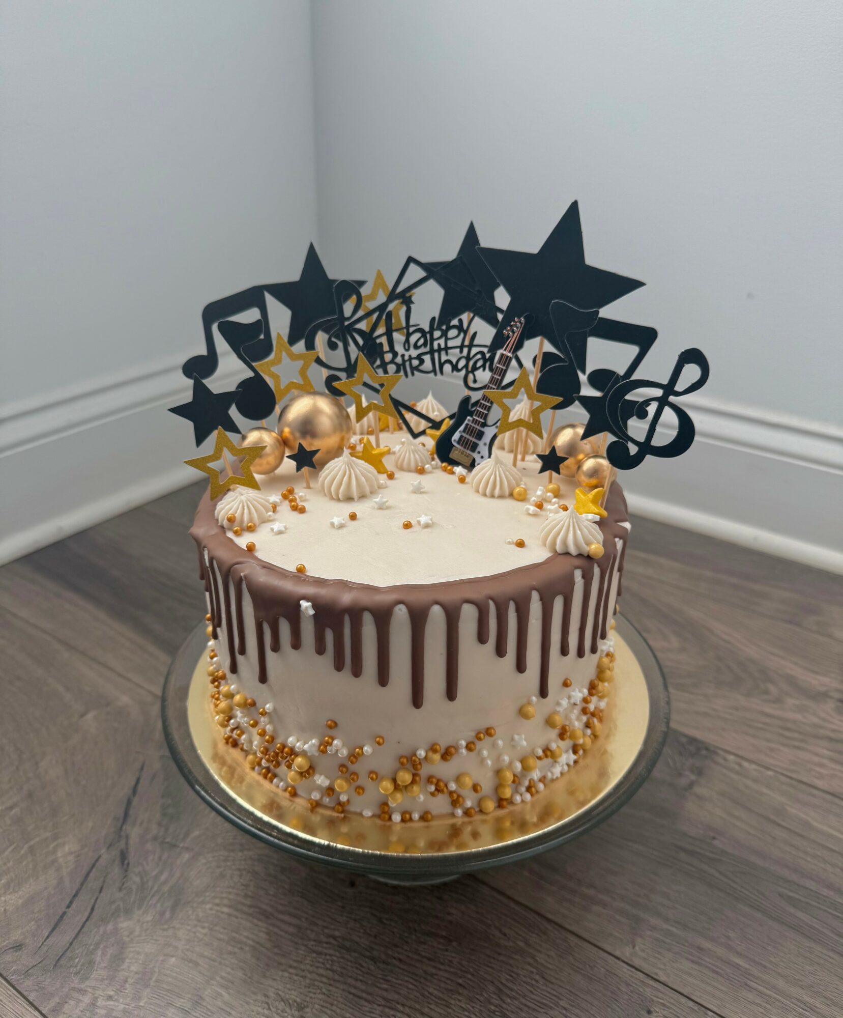 Round birthday cake with white frosting, gold and white decorations, and black musical note and star toppers, on a silver tray.