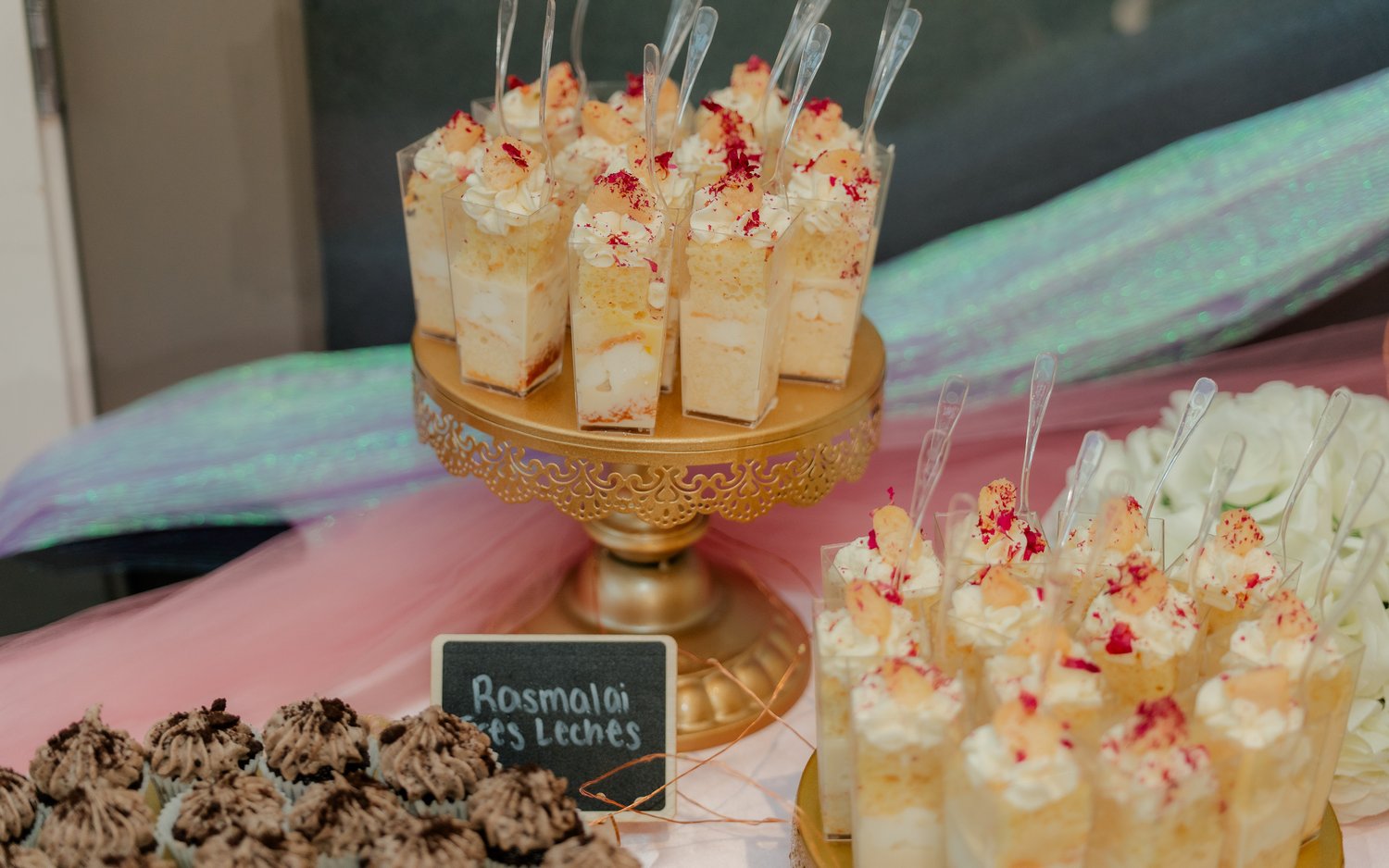 Dessert display with small cake slices topped with cream and red sprinkles, arranged on a gold stand, with other treats nearby.