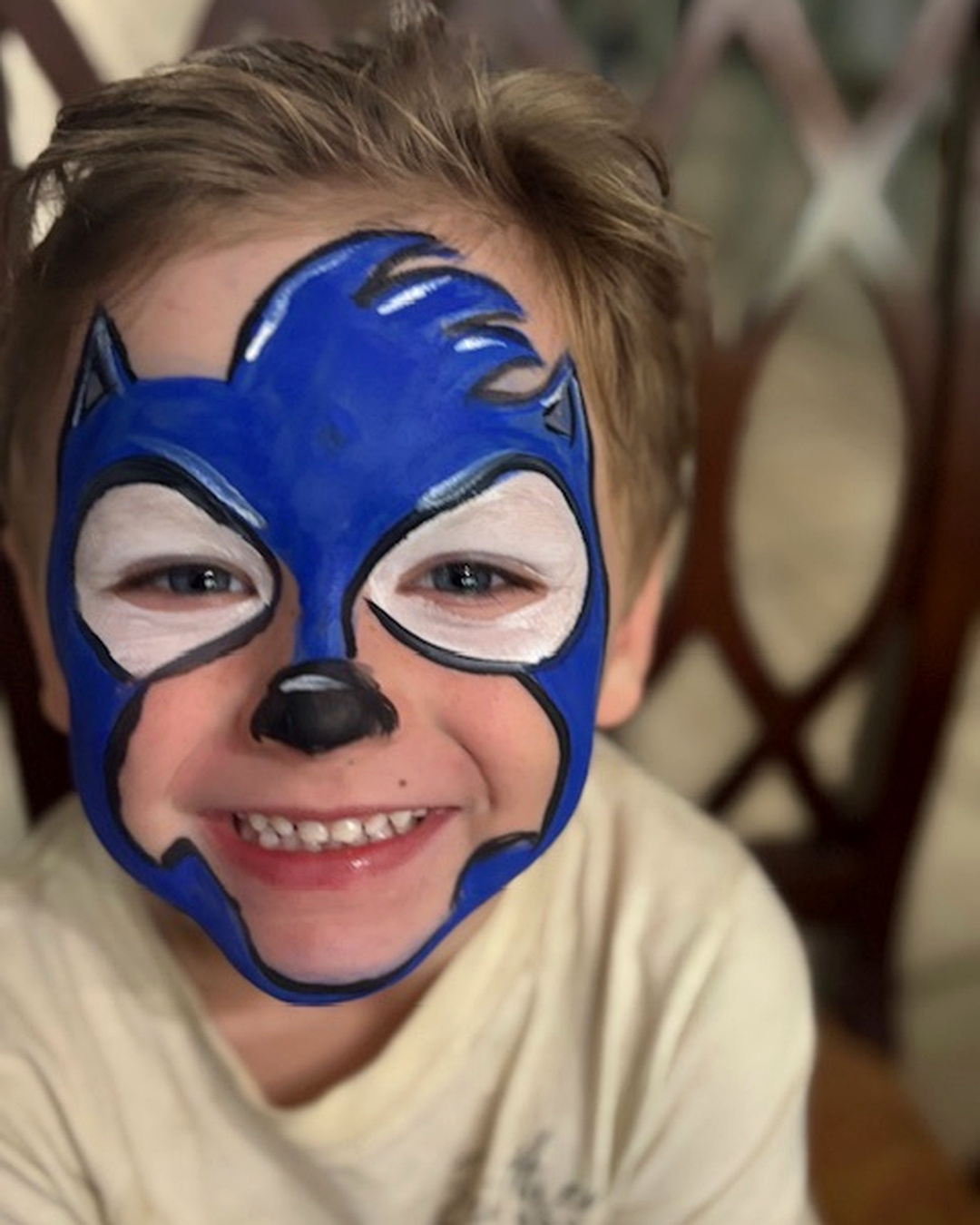 Child with face painted as Sonic the Hedgehog, smiling, indoors with a wooden chair in the background.
