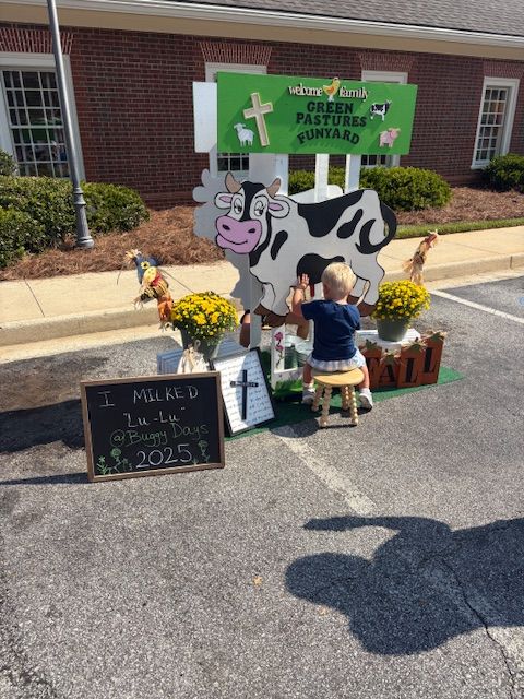 People gather around a display with a cow sculpture and a chalkboard sign, outside a brick building with windows.