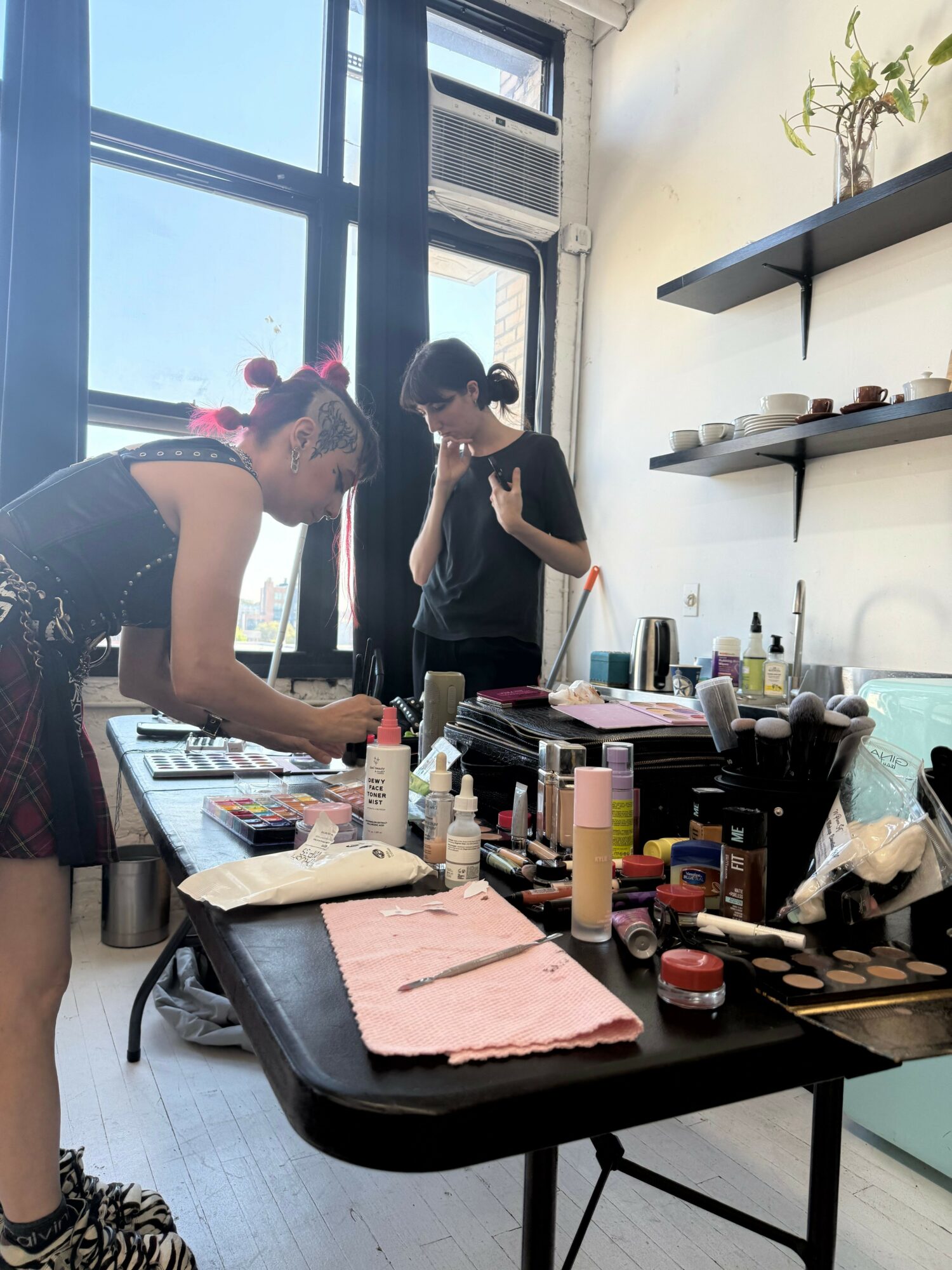 Two women in a room with large windows, one sitting at a table with makeup products, the other standing nearby.