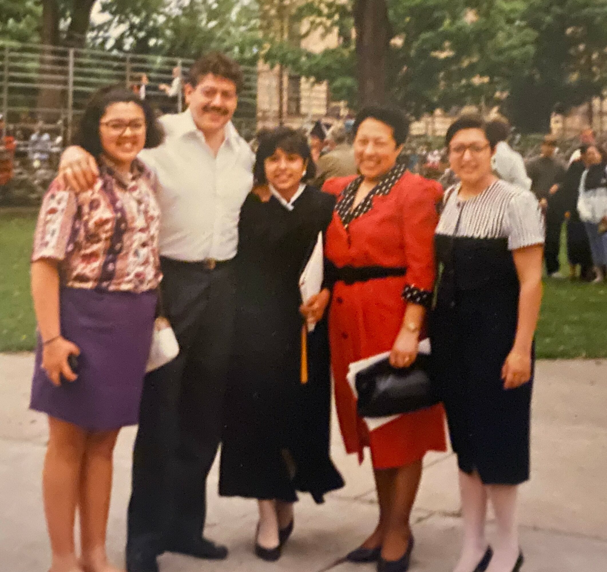 Group of five people standing outdoors, smiling, with trees and a park in the background.