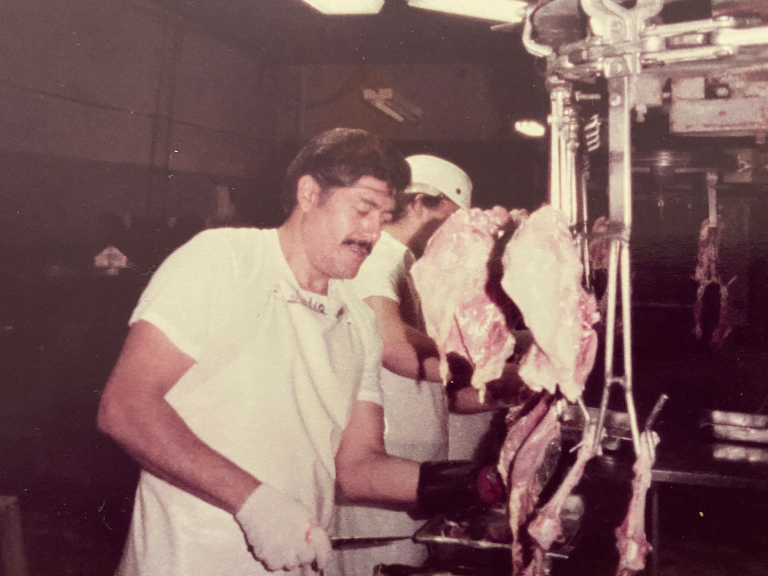 Person in white shirt and gloves working with meat on a hanging scale in a butcher shop.