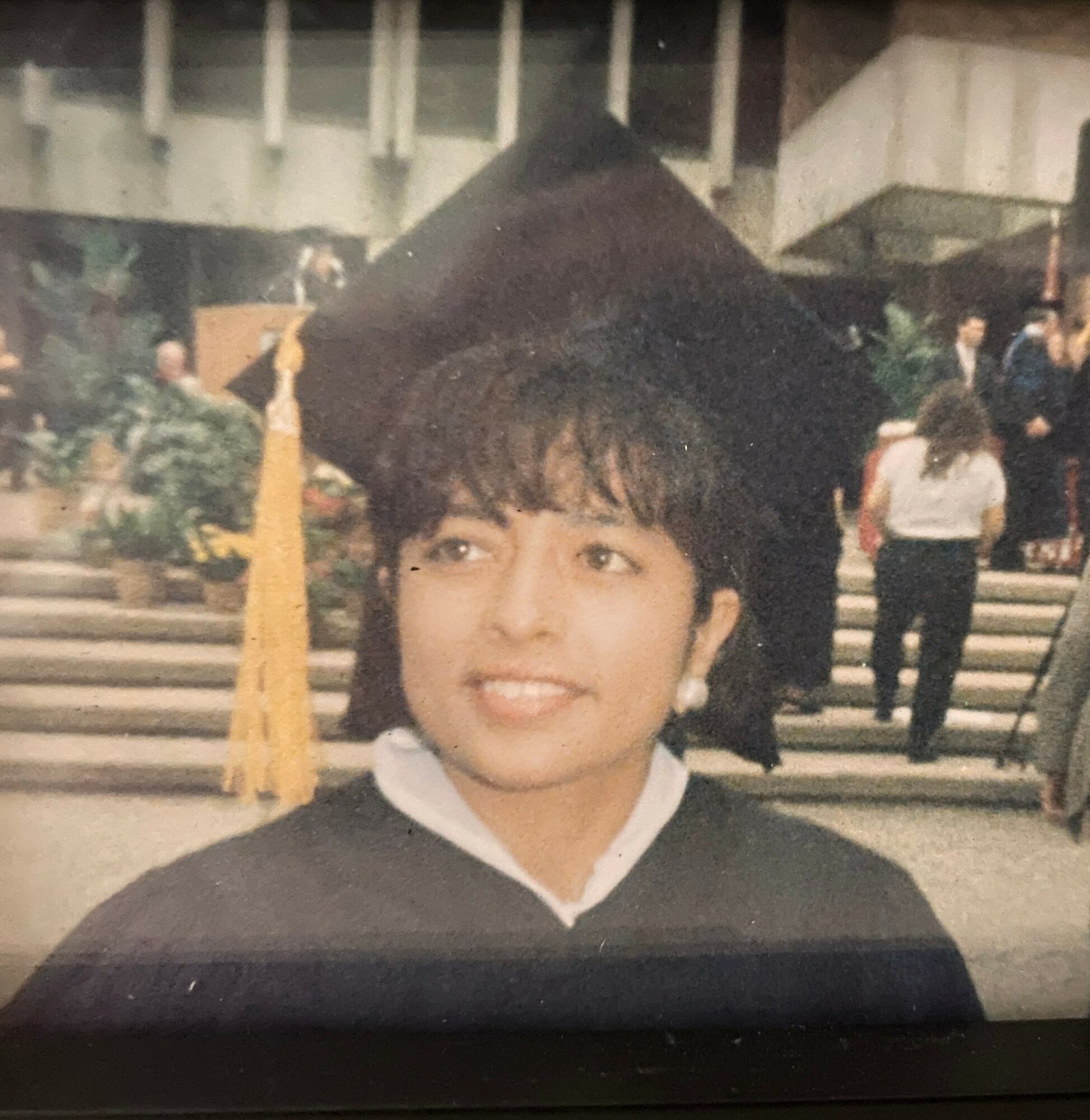 Person in graduation cap and gown smiling outdoors, with stairs and people in background.