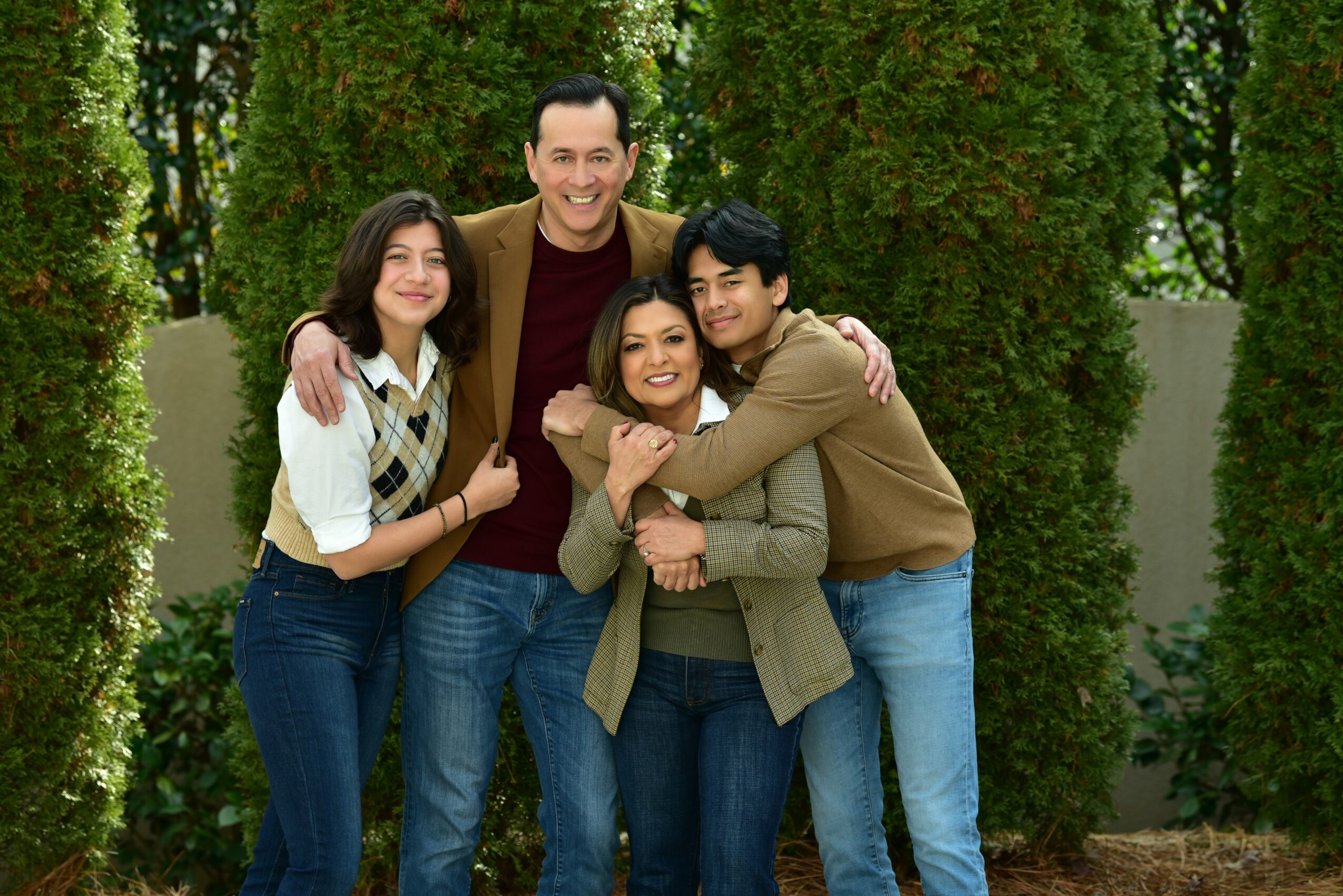 Group of four people smiling and hugging outdoors with green trees in background.