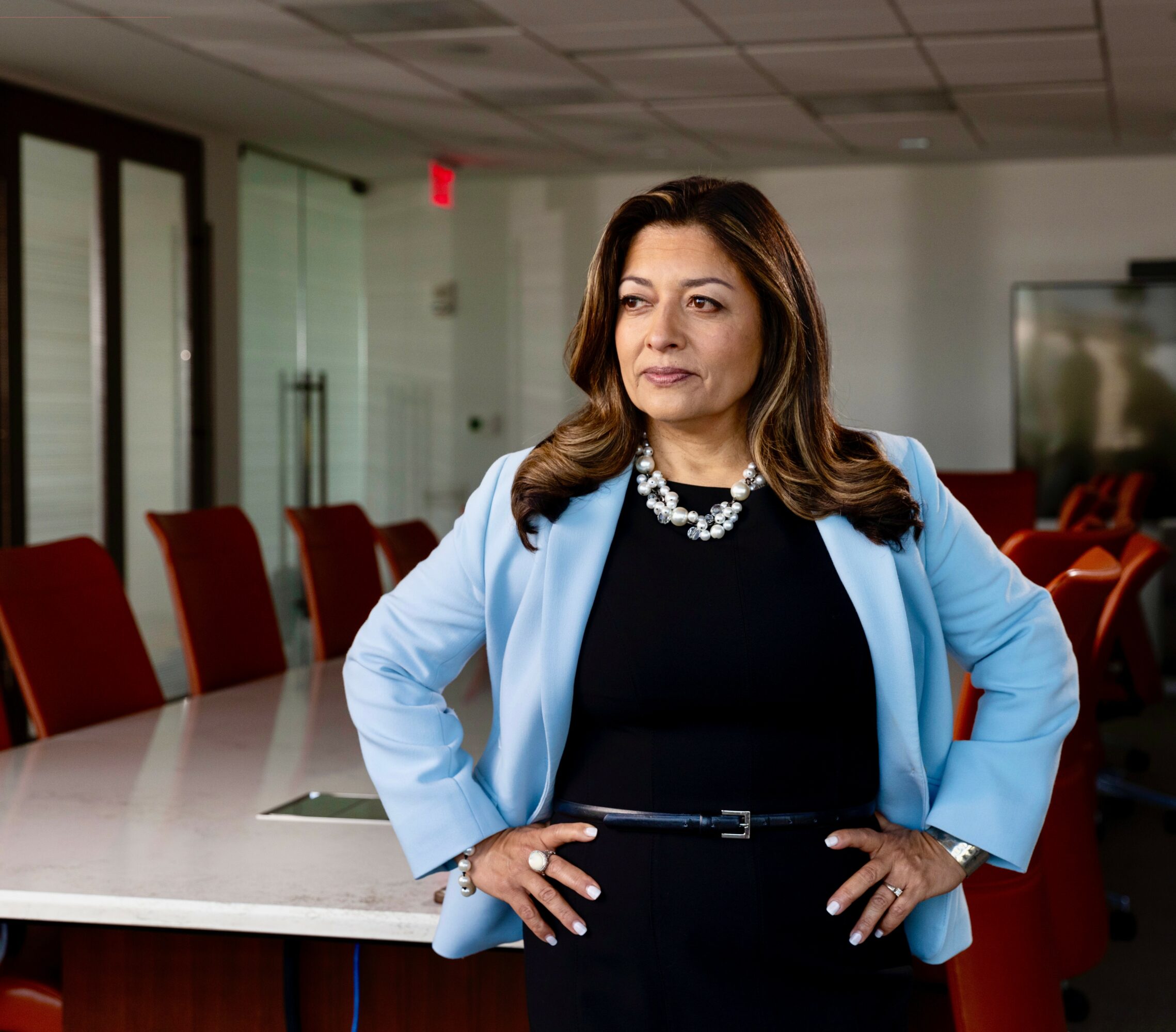 Woman in a light blue blazer and black dress stands in a conference room with red chairs and glass doors.