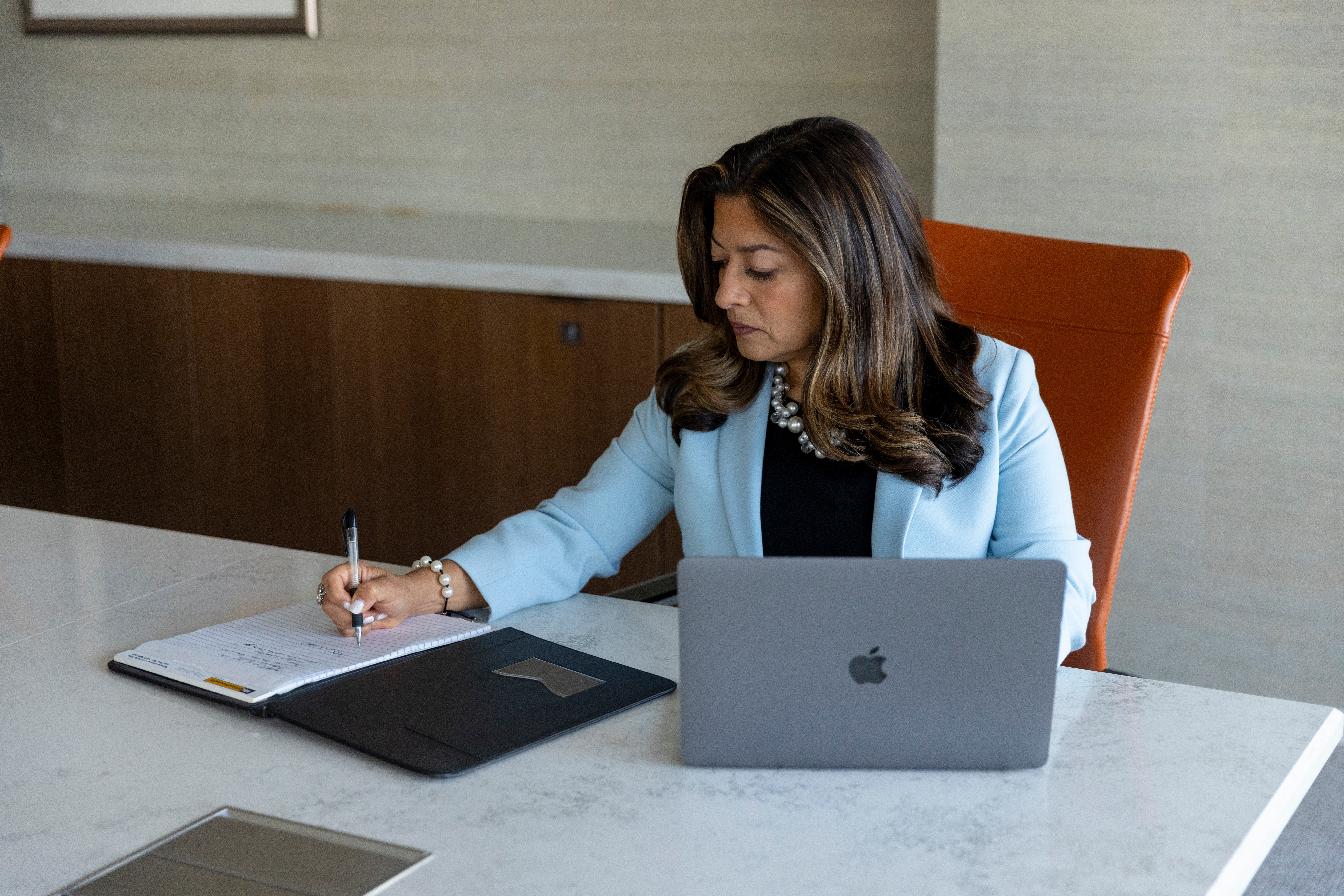 Woman sitting at a desk with a laptop, writing in a notebook, in a professional setting.