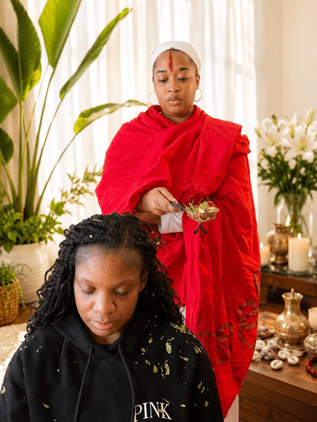 Two women indoors, one in a red shawl holding a spoon, the other with braided hair looking down, surrounded by plants and flowers.