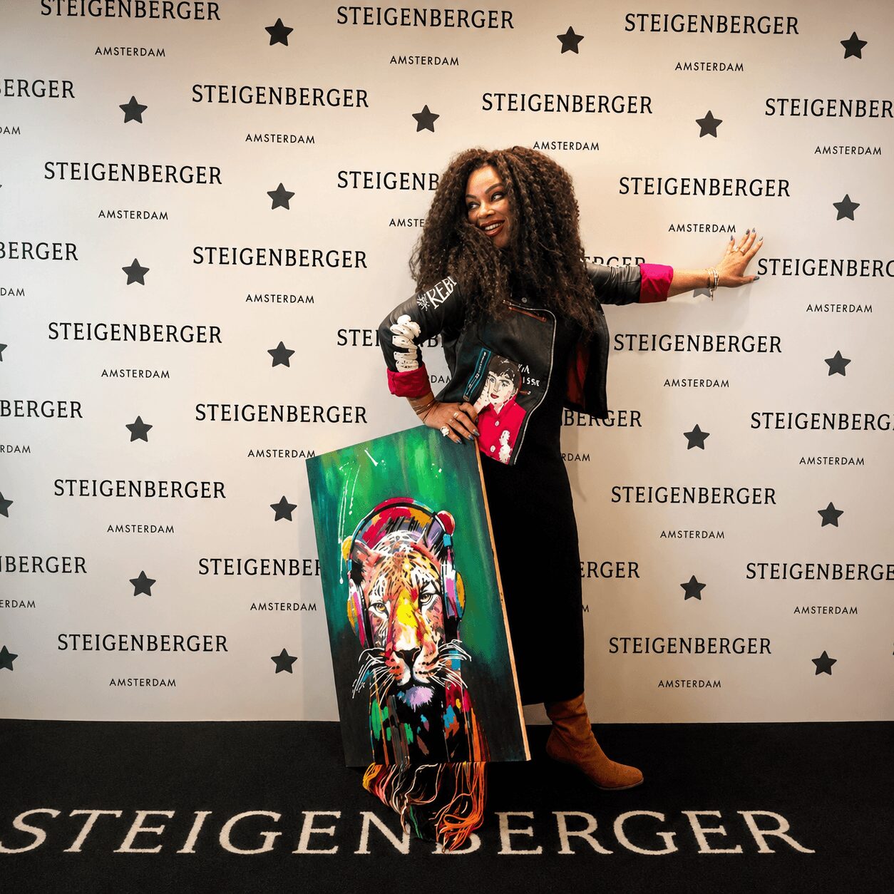 Woman with curly hair poses with a colorful painting in front of a branded backdrop, smiling and pointing.