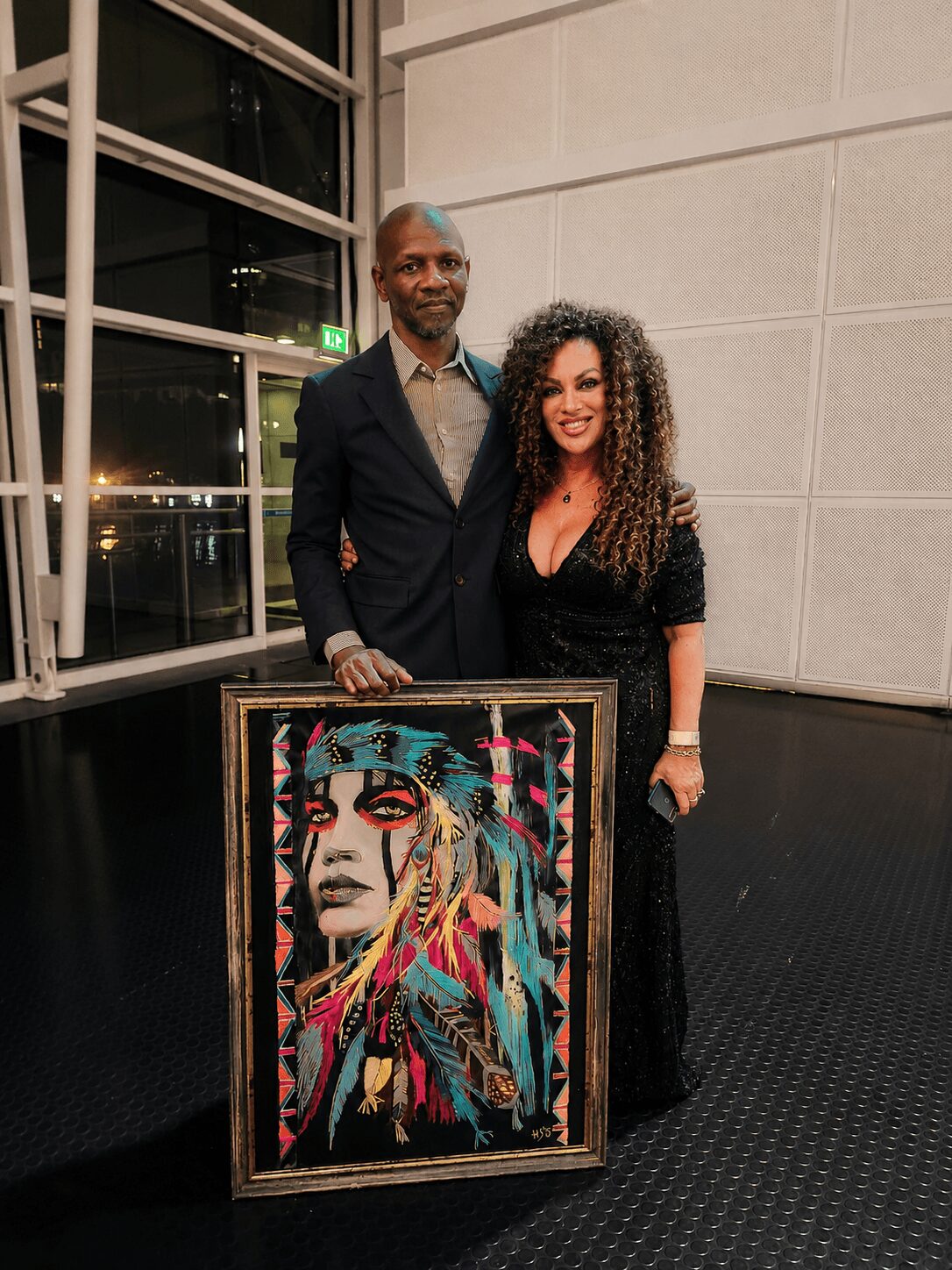 Two people stand behind a colorful portrait painting in an indoor setting, with large windows and a white wall in the background.