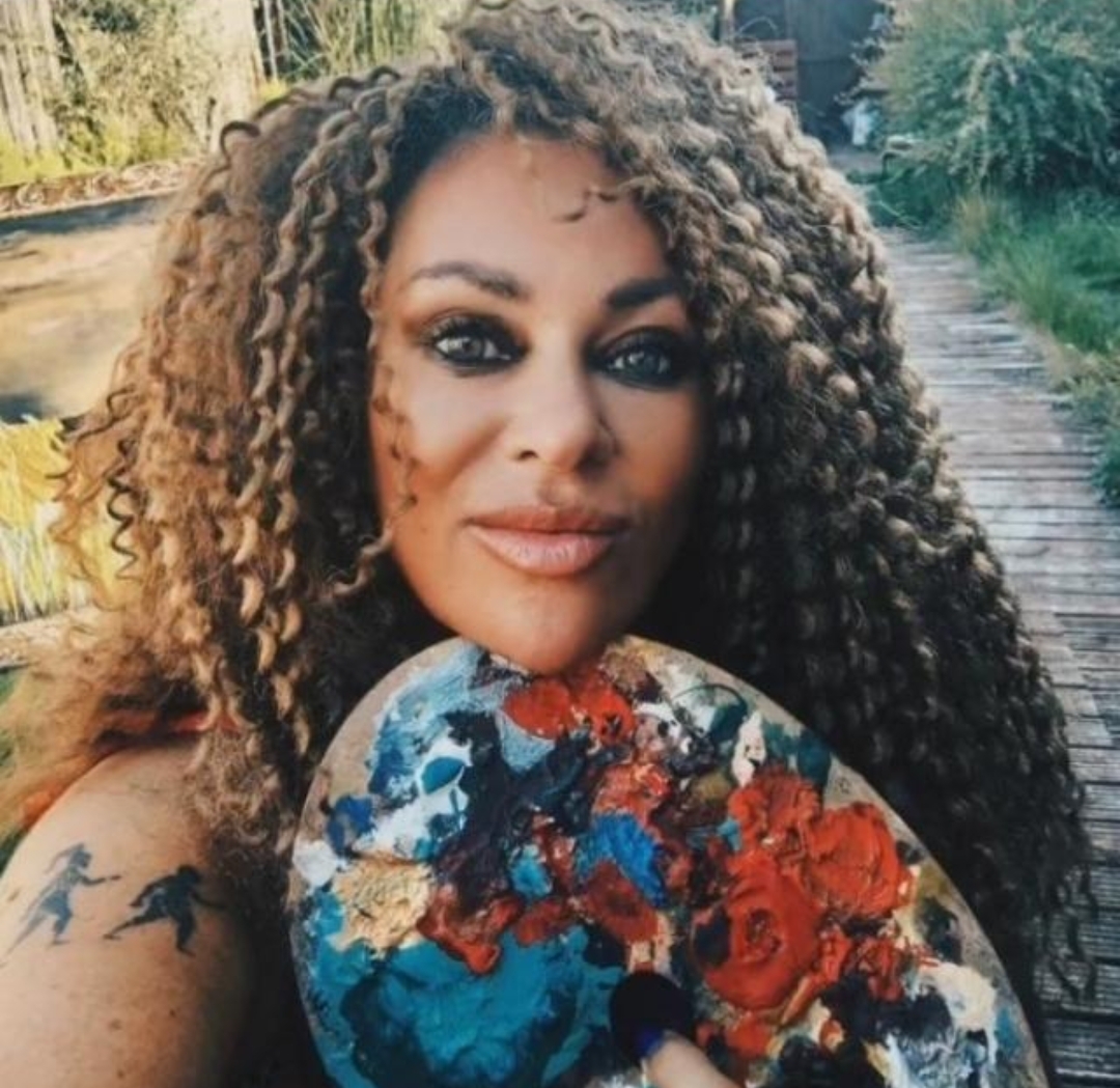 Woman with curly hair holding a colorful floral fan outdoors, smiling, with trees and a wooden path in the background.