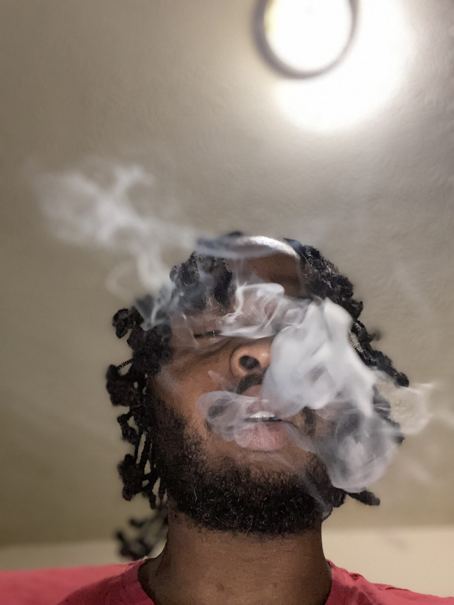 Person with curly hair and beard exhales smoke, partially obscuring face, ceiling light visible above, neutral background.