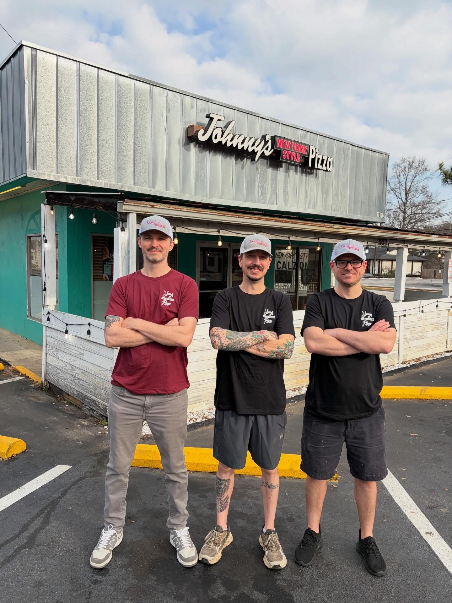 Three men stand outside a restaurant with crossed arms, wearing casual clothes and caps, in a parking lot.