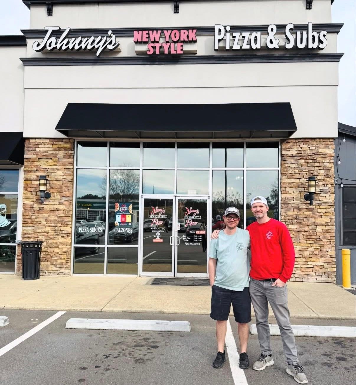 Two people stand outside Johnny's Pizza & Subs restaurant with glass doors and brick walls, smiling at the camera.