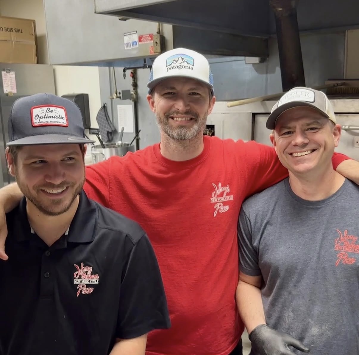 Three smiling people in a kitchen, two men wearing caps and a woman with a gray shirt, all with arms around each other.