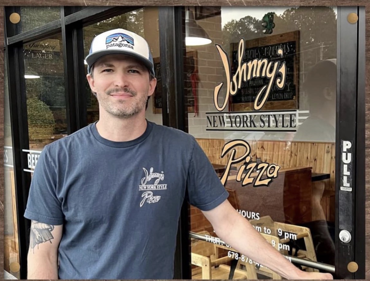 Man wearing a blue shirt and cap standing outside restaurant with glass door, wooden interior, and signs for pizza and New York style.