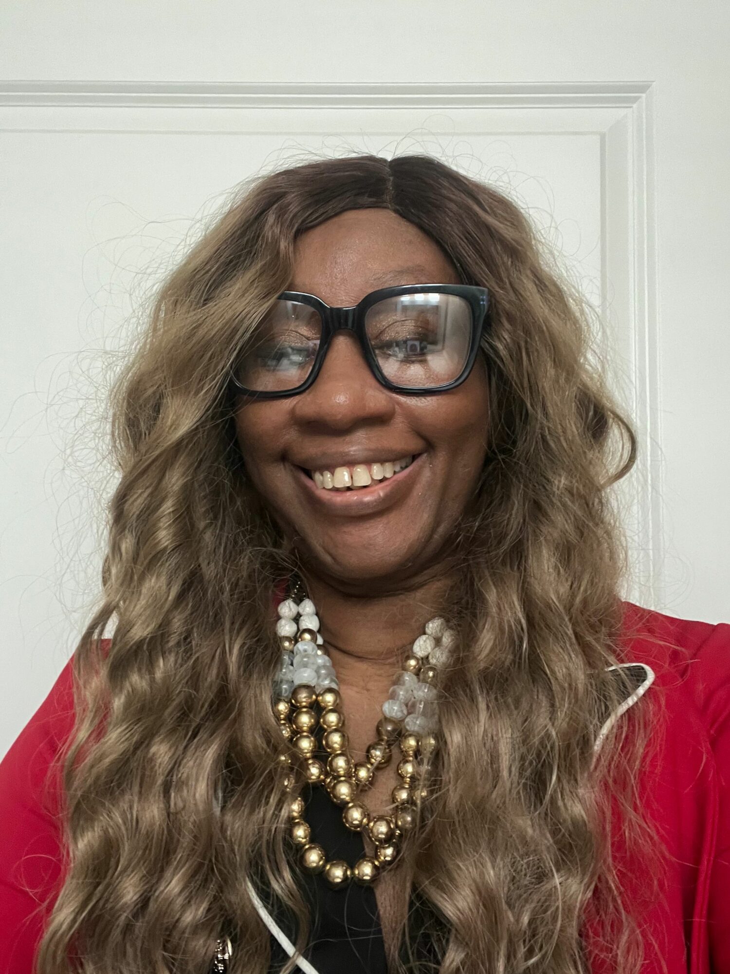Woman with long wavy hair, glasses, smiling, wearing a red top and layered pearl and gold necklace.