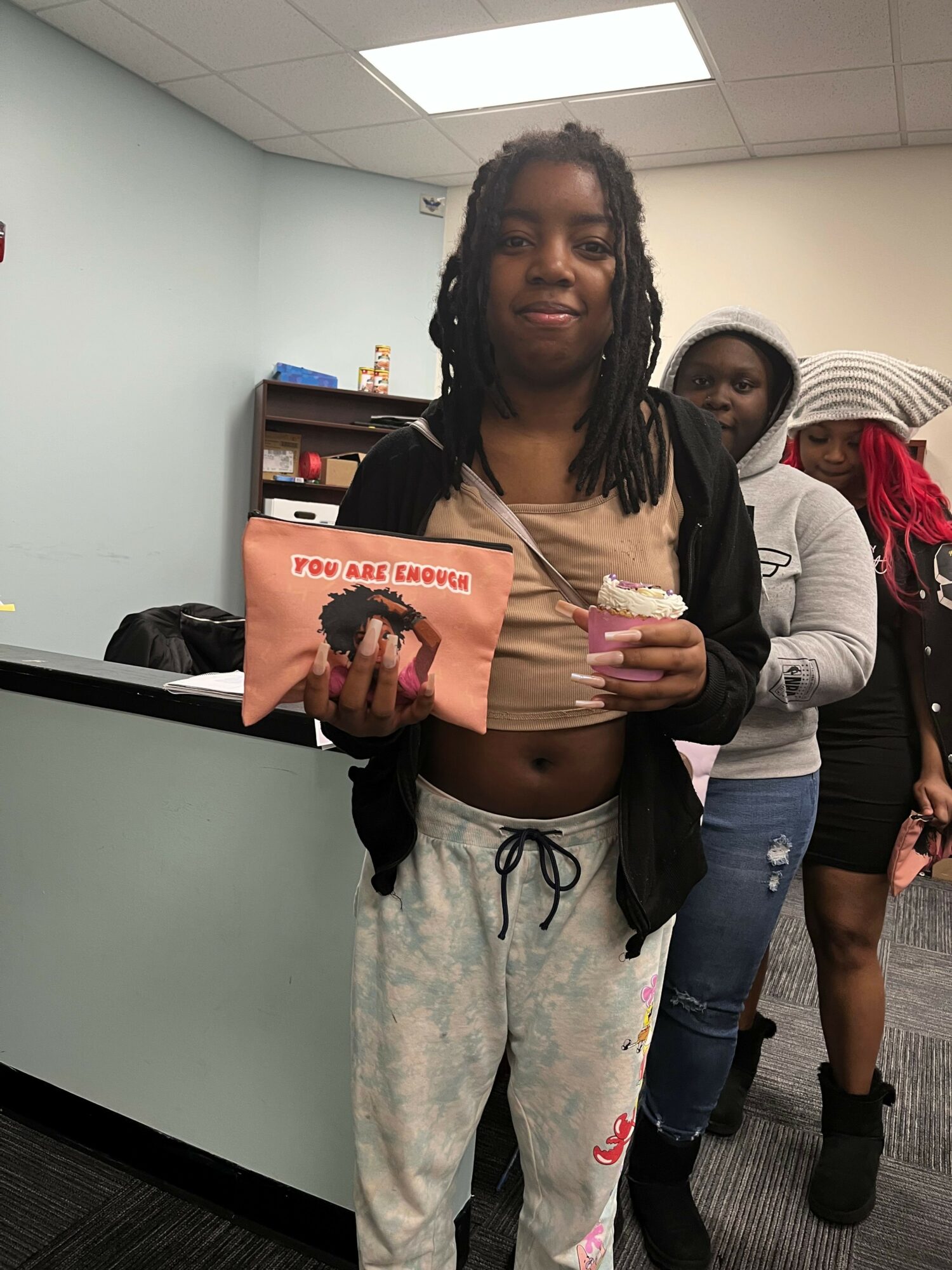Person with long dreadlocks holding a pink bag, standing in an office with a white wall and bookshelf.
