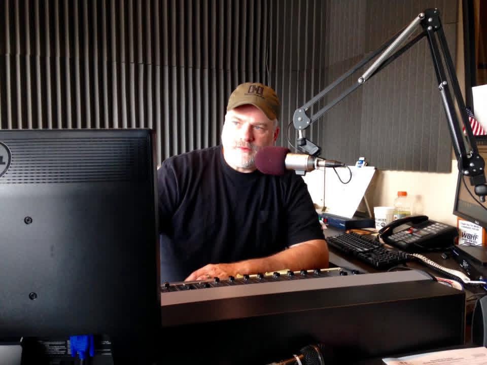 Man with a beard and cap sitting at a radio station desk with microphone and computer equipment.