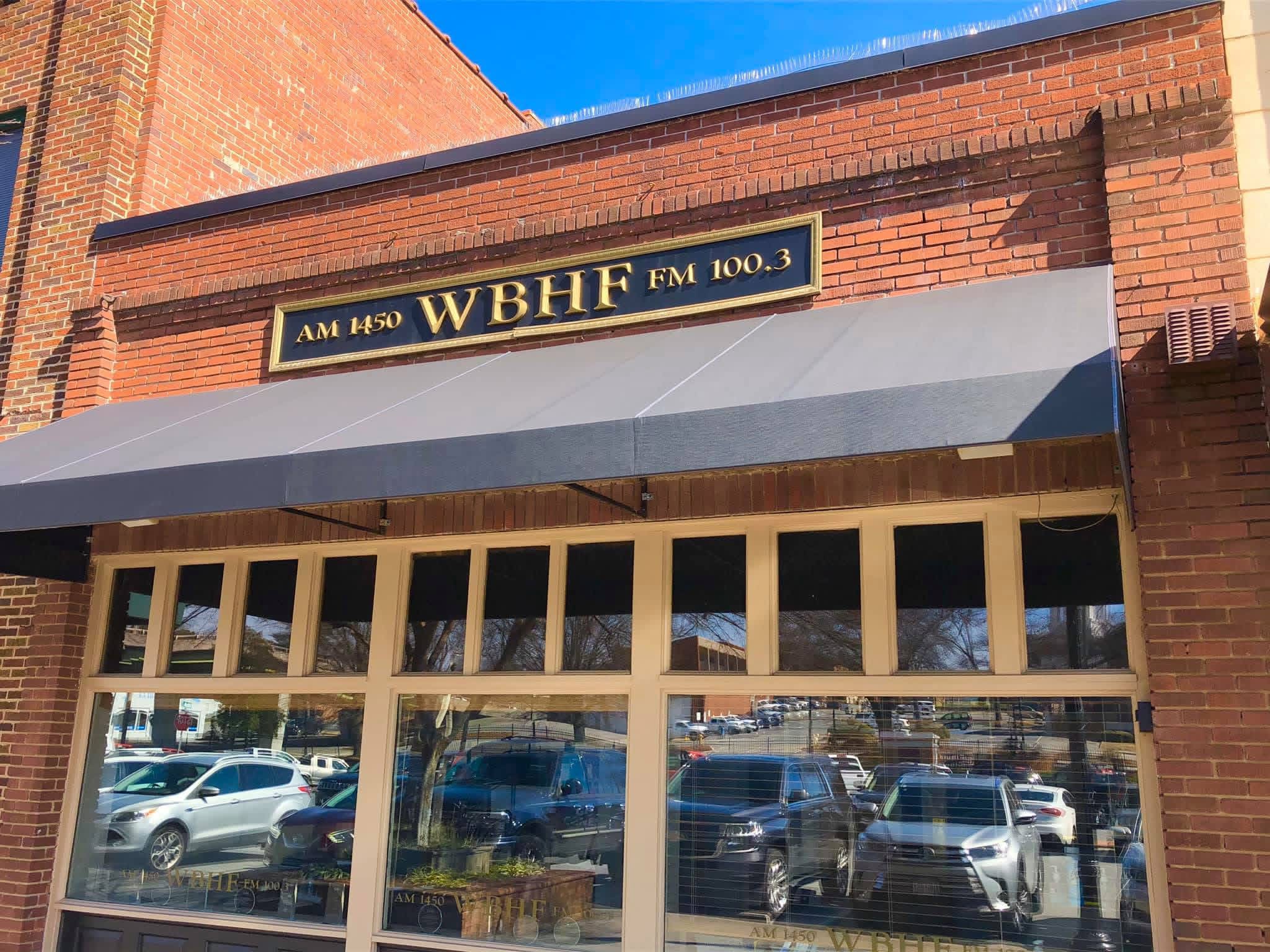 Storefront with a sign reading 'WBHF FM 100.3' and large windows reflecting parked cars outside.