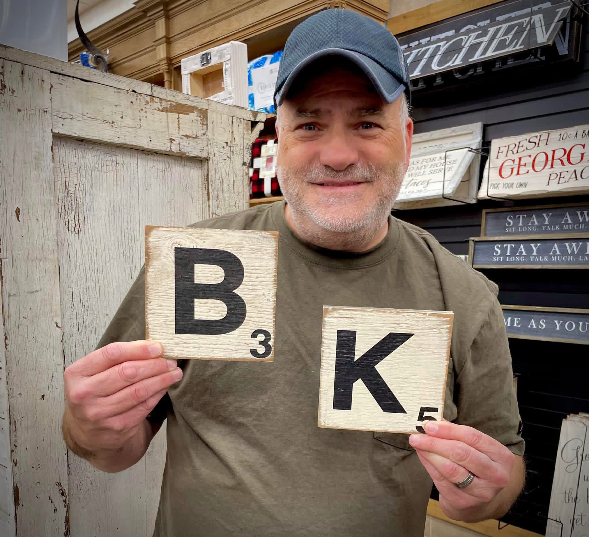Man holding Scrabble tiles B and K in front of a store display, smiling, wearing a cap and casual clothing.