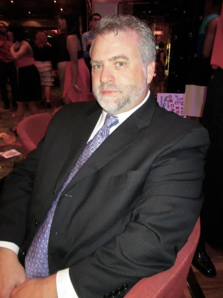 Man in a suit sitting at a table in a dimly lit indoor setting with people in the background.