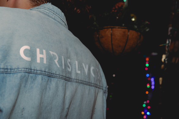 Back of a person wearing a light blue denim jacket with the name 'CHRISLVC' printed on it, nighttime setting with colorful lights in the background.