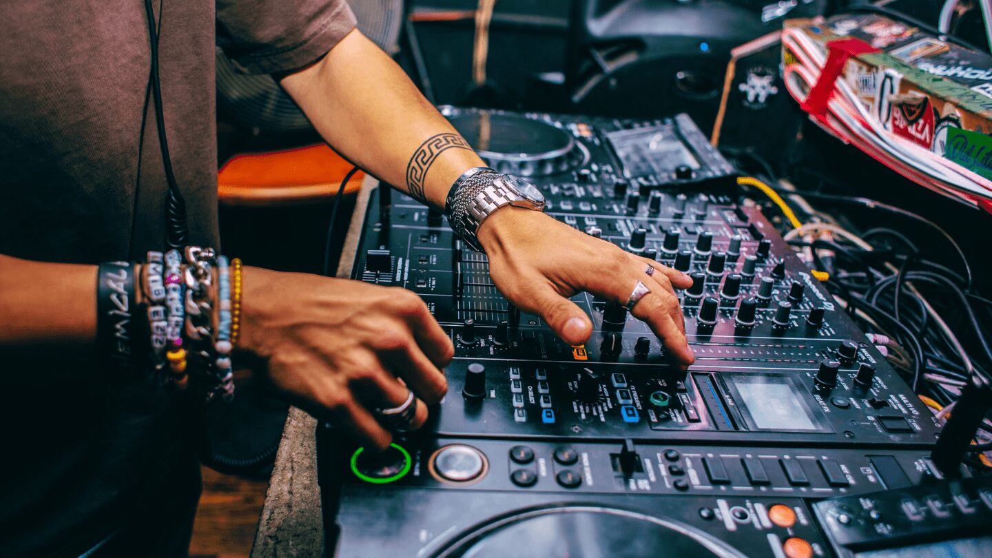 Person's hands adjusting DJ equipment with colorful bracelets, mixing music at a DJ booth, with various controls and cables visible.
