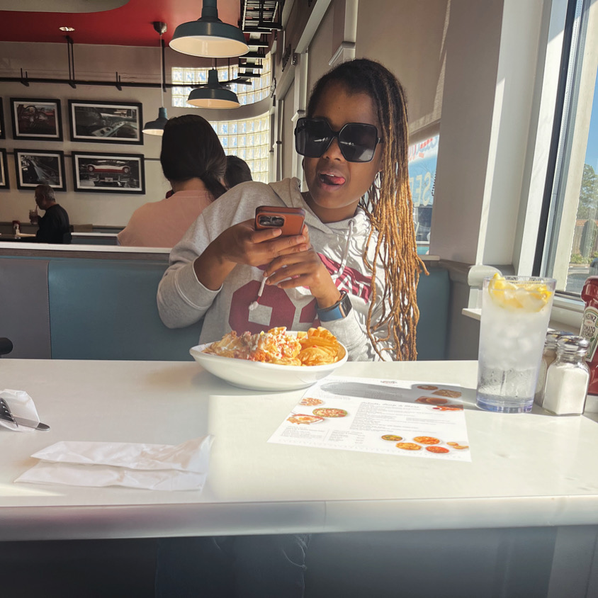 Woman with sunglasses using a phone at a restaurant table with food and drink, large window in background