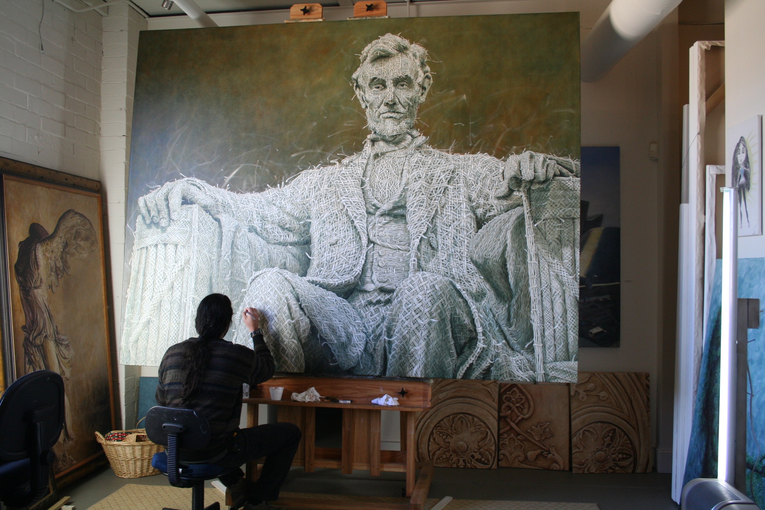 Person sitting in front of a large portrait of a seated man with arms outstretched, in an indoor setting.