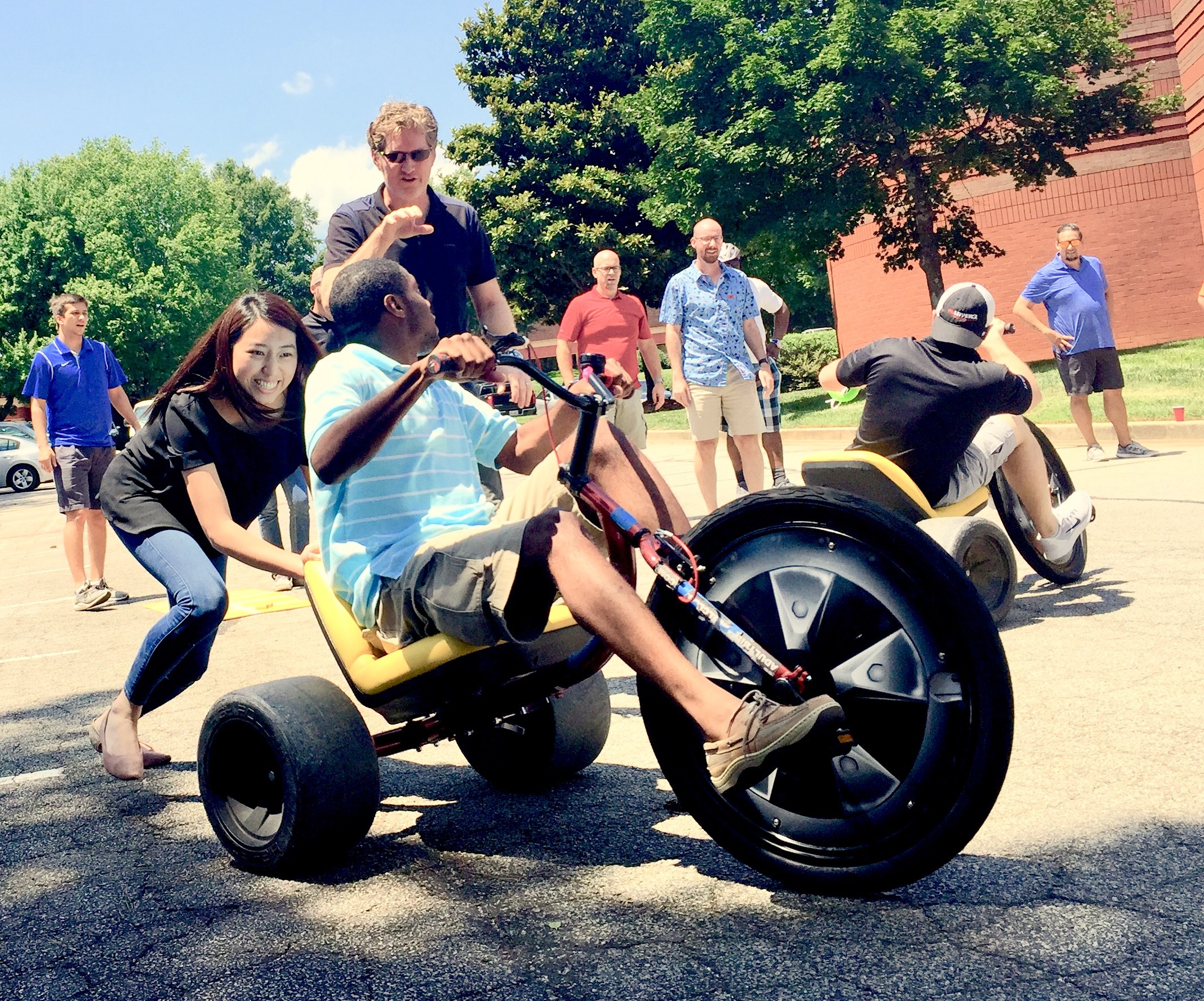 Person riding a yellow pedal-powered vehicle with a large front wheel, surrounded by people outdoors on a sunny day.