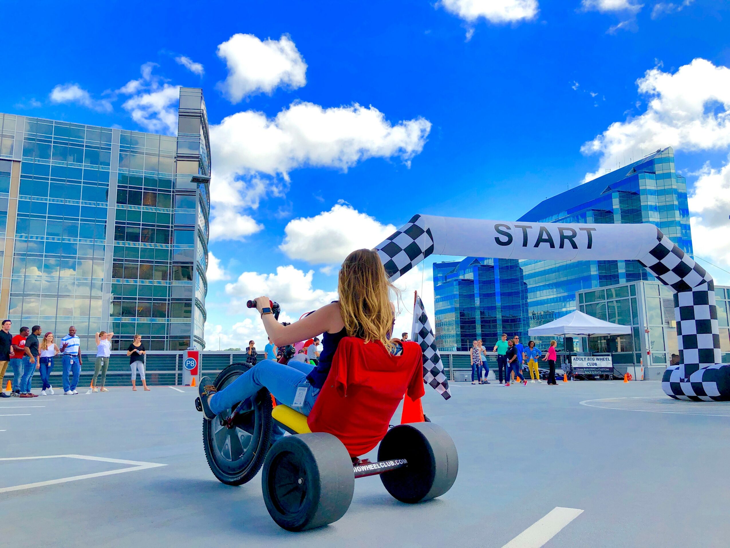 A woman riding a tricycle with a red seat in an outdoor parking lot, starting a race under a checkered start banner, with modern glass buildings and a blue sky with clouds in the background.